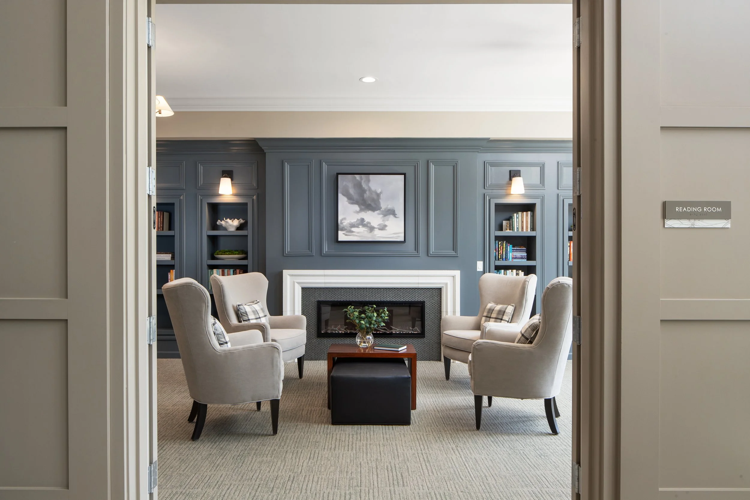 A cozy reading room with beige armchairs, a fireplace with a white mantel, blue built-in bookshelves, and a painting of clouds above the fireplace. There are pillows on the chairs and a small table with a plant and books in front.