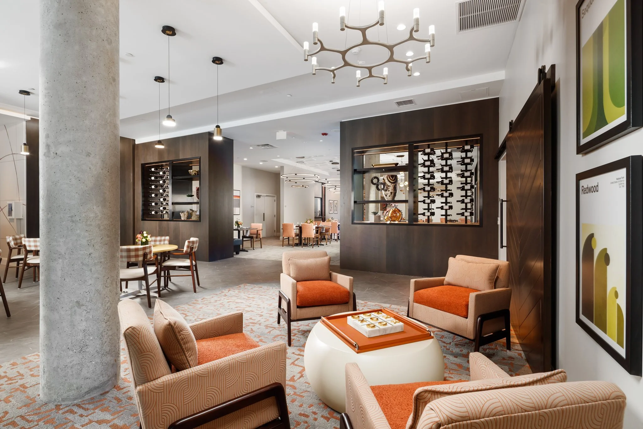 Interior of a modern hotel lounge featuring beige armchairs, a white round coffee table with decorative items, and a large gray concrete column. The lounge has contemporary lighting fixtures, including a chandelier and pendant lights, with dark wood paneling on the walls and a wine rack in the background.