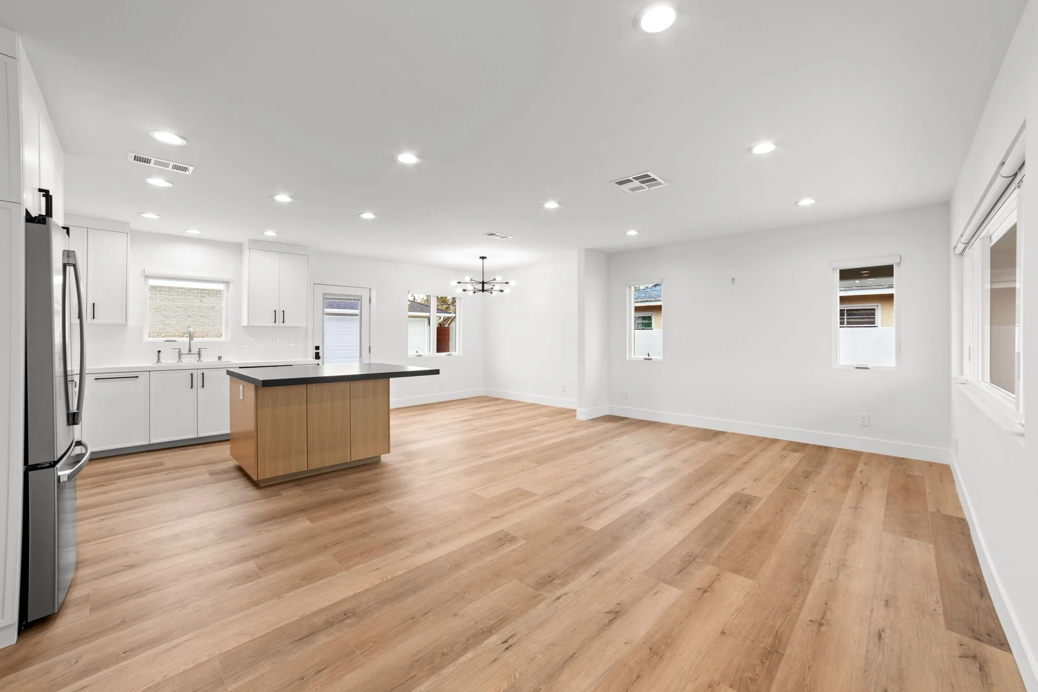 Empty modern kitchen with white cabinets, wooden island, wood flooring, multiple windows, and recessed ceiling lights.