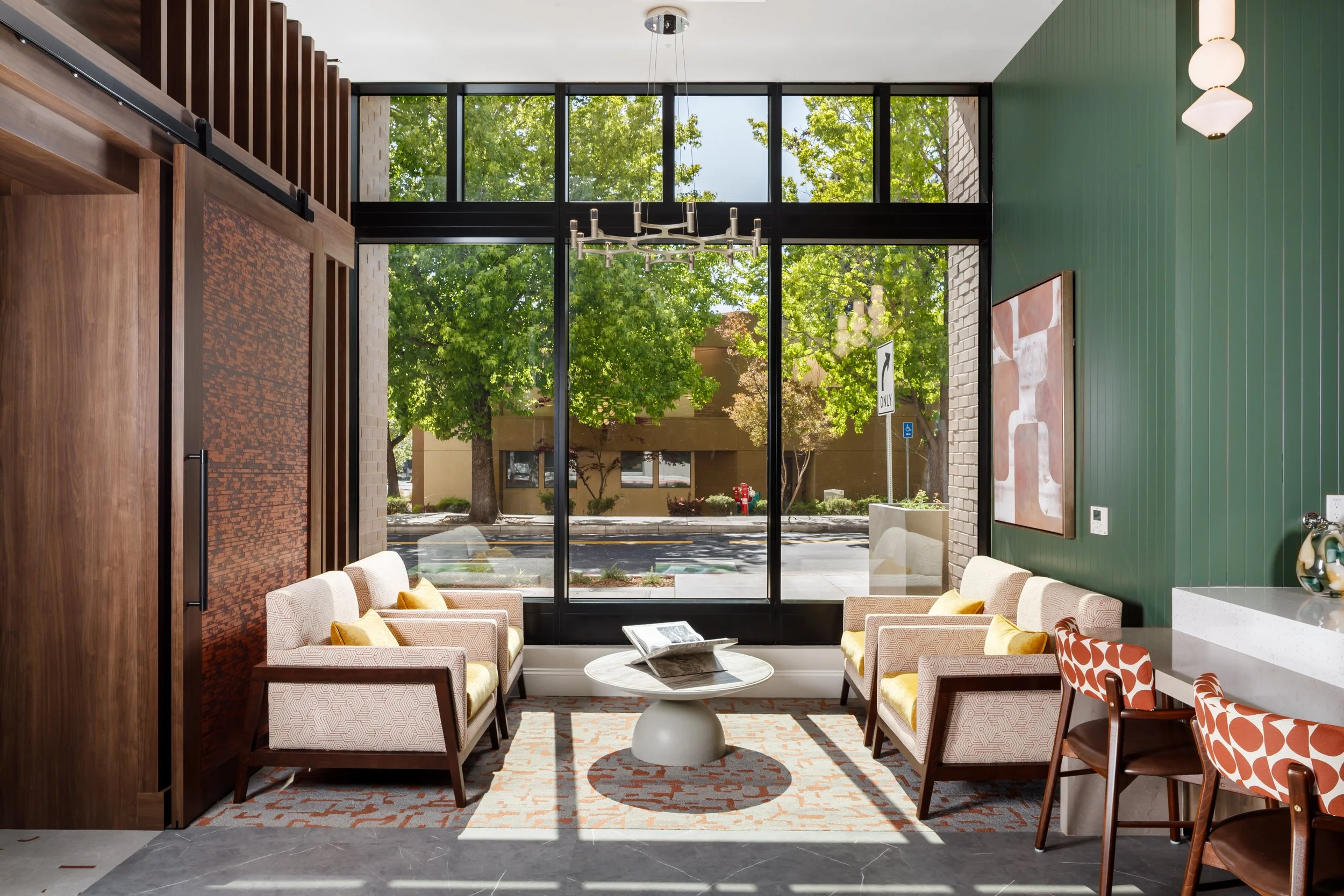 Modern interior space with large front window, four beige armchairs with yellow pillows, a round white marble coffee table with a magazine, artwork on green wall, patterned orange and white chairs, and outside view of trees and street.