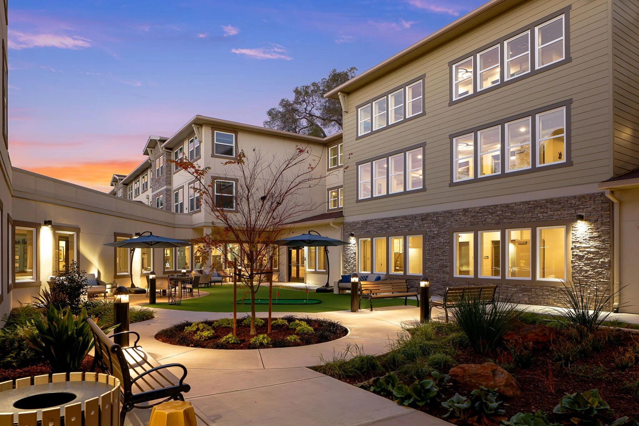 Apartment complex courtyard at sunset with outdoor seating, lighting, trees, and multiple windows in the buildings.