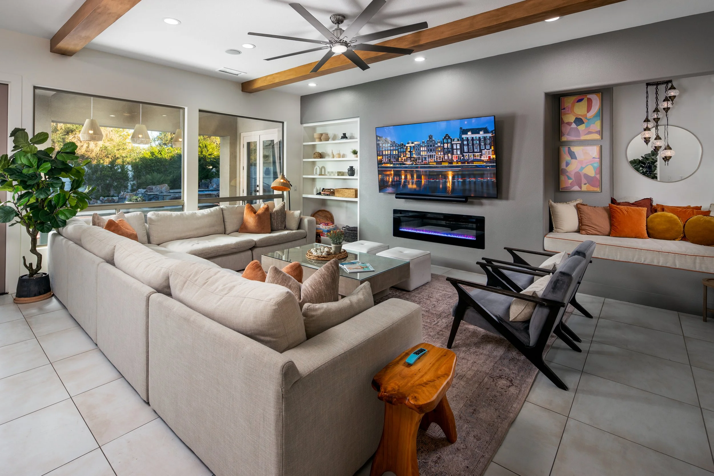 Modern living room with large L-shaped beige sofa, wooden coffee table, black rocking chairs, a wall-mounted flat-screen TV above a fireplace, built-in white shelves, a big potted plant, and large windows showing greenery outside.