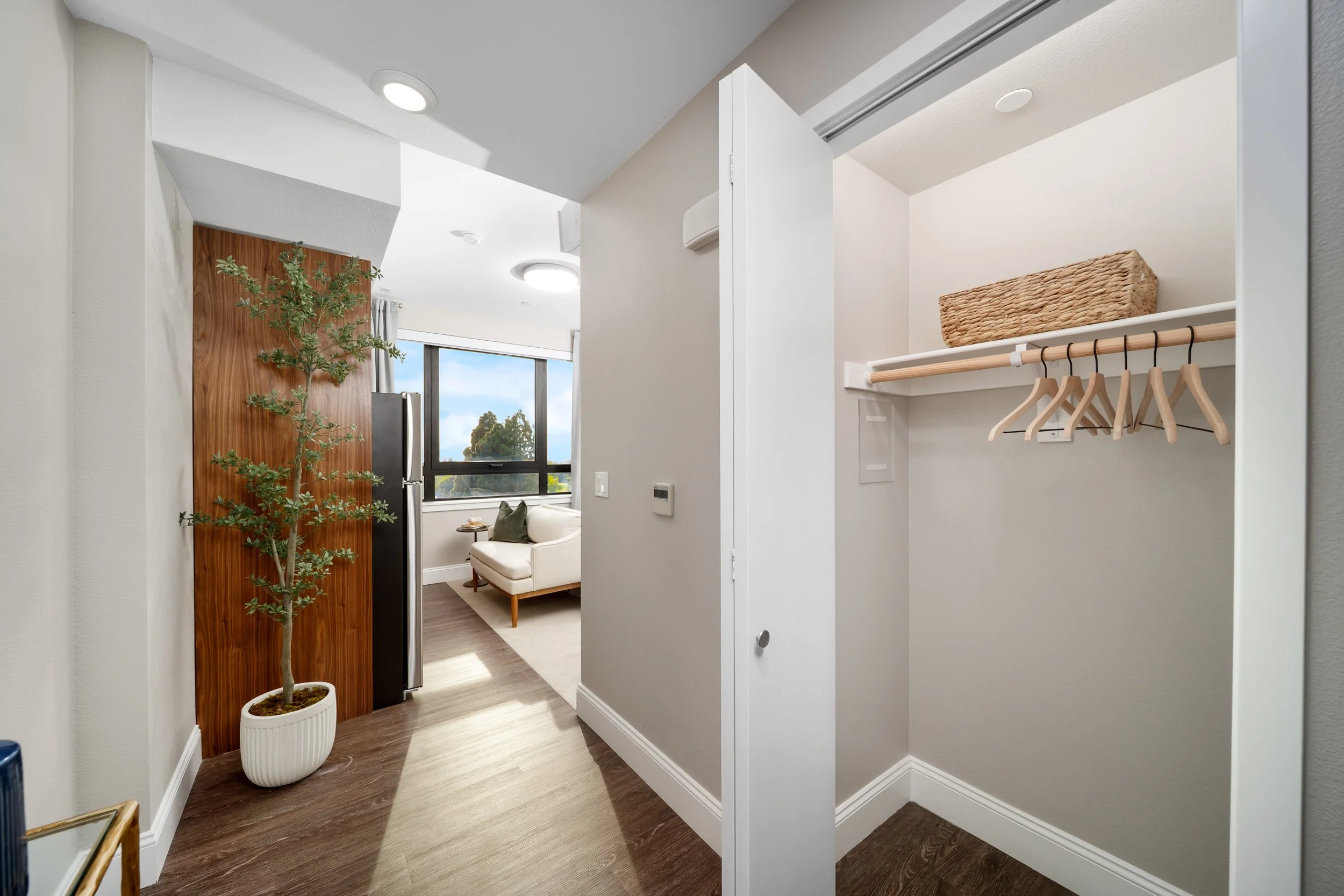 An interior view of an apartment entryway and living room, showing a partially open closet with a wicker basket and wooden hangers on a shelf, a wall-mounted thermostat, and a living room with a window, white couch, and a hardwood floor.