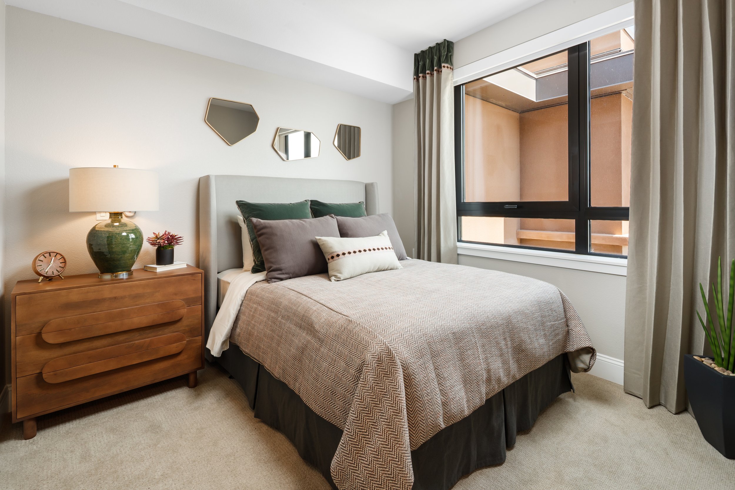 A cozy bedroom with a bed, decorative pillows, a wooden nightstand, a lamp, a small clock, a potted plant, and wall mirrors near a large window with curtains.
