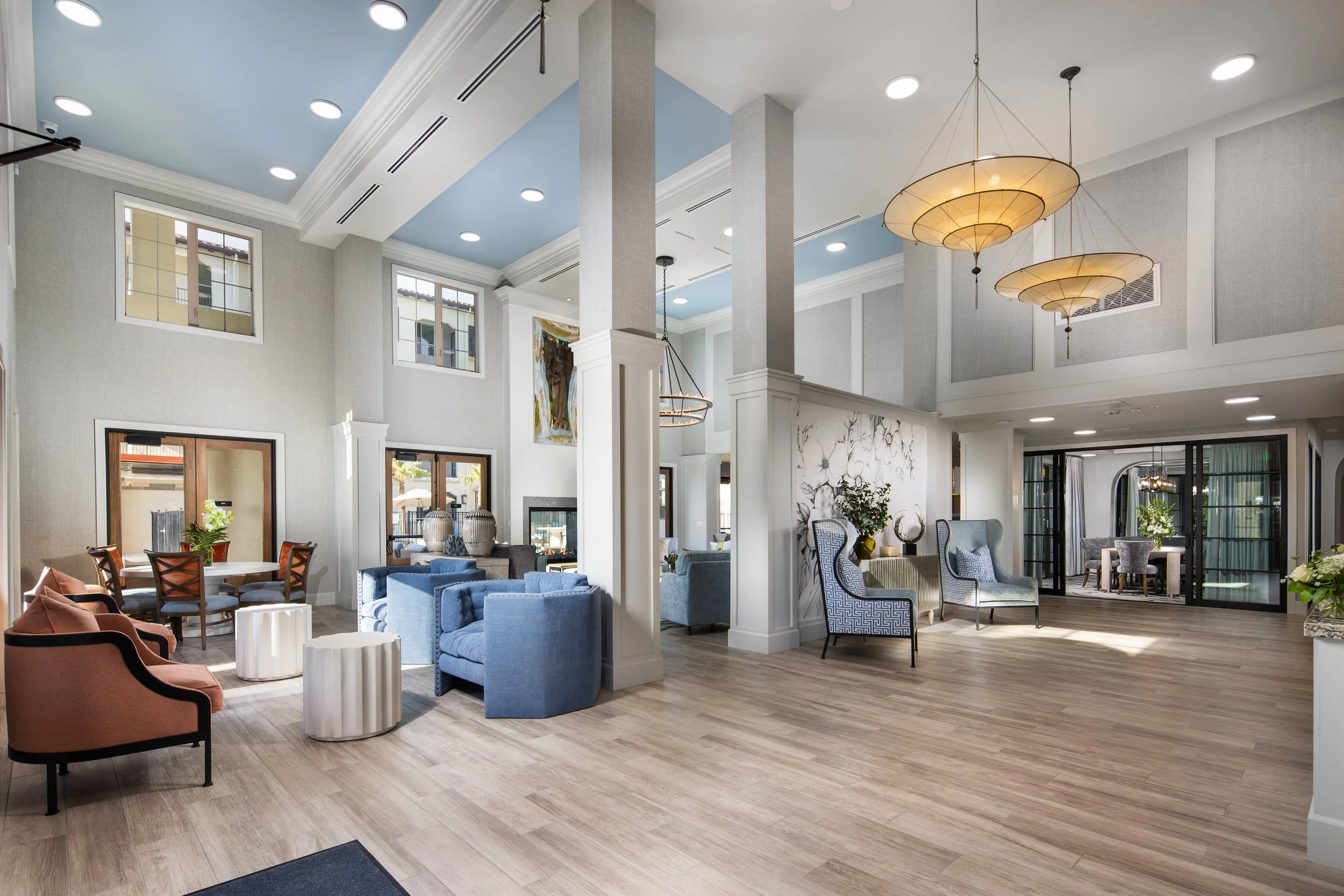 Interior of a spacious, modern lobby with high ceilings, light-colored walls, and large windows. It features various seating options including blue armchairs, pink armchairs, and patterned high-back chairs. There are decorative pendant lights hanging