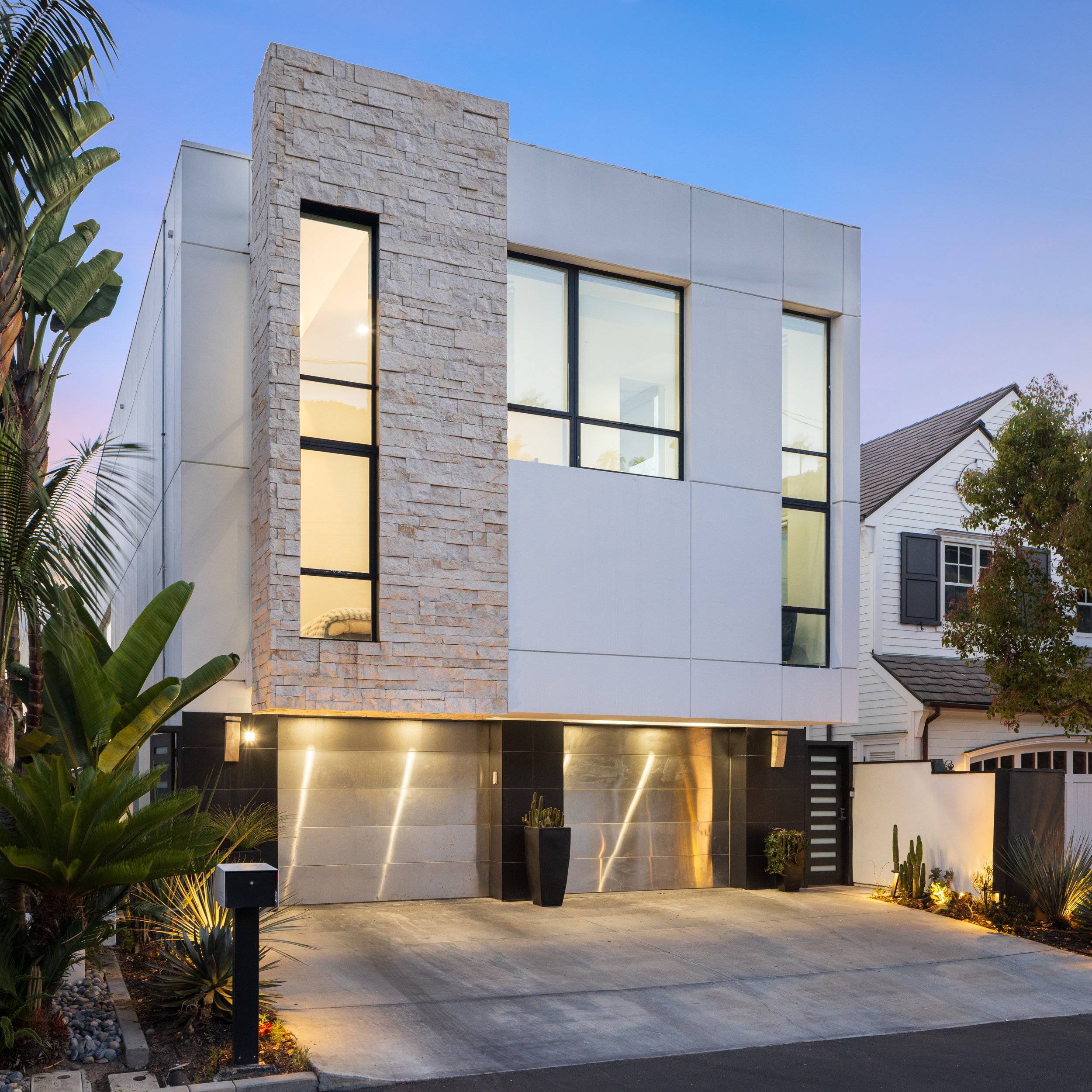 Modern house with stone and white exterior, large windows, and landscaped front yard with plants and lighting at dusk.