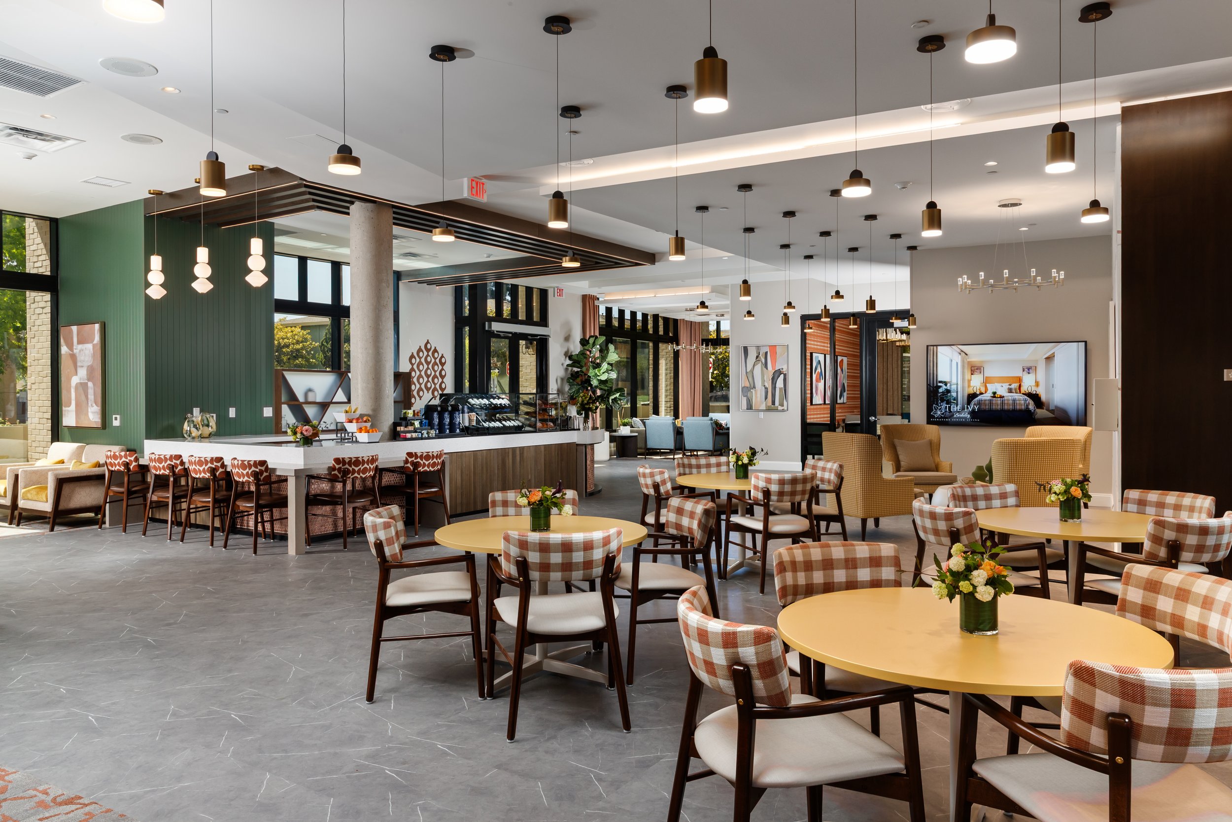 Interior of a modern cafe with round tables, floral centerpieces, checkered and solid chairs, a bar area, large windows, artwork on the walls, and pendant lighting.