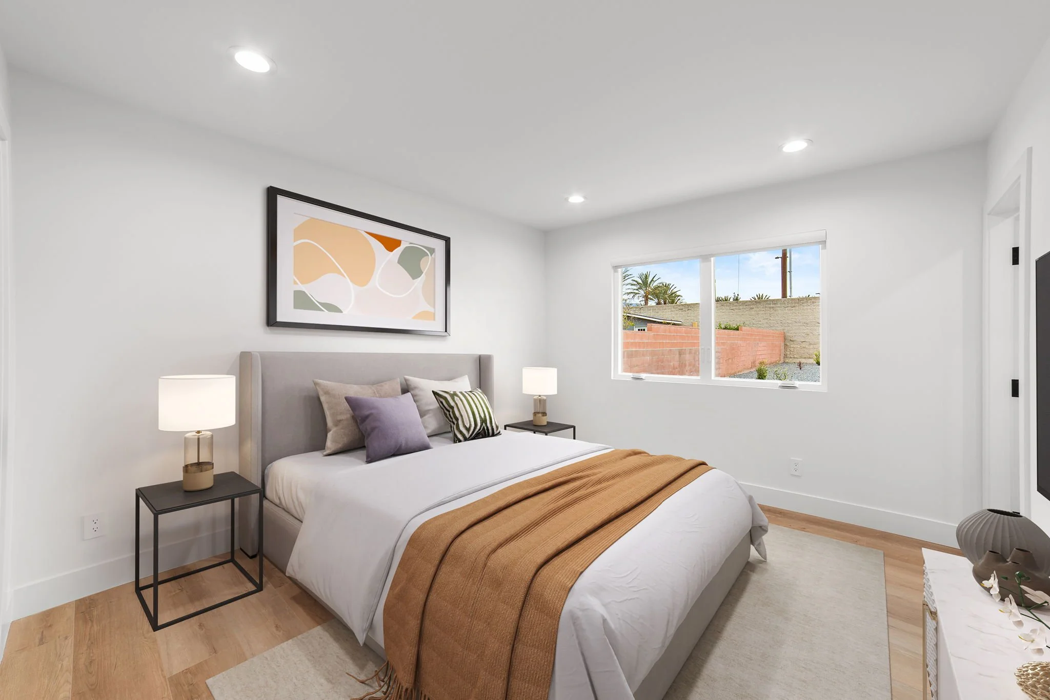 A bedroom with a bed, pillows, two bedside tables with lamps, a large window showing palm trees and a brick wall outside, and artwork on the wall above the bed.