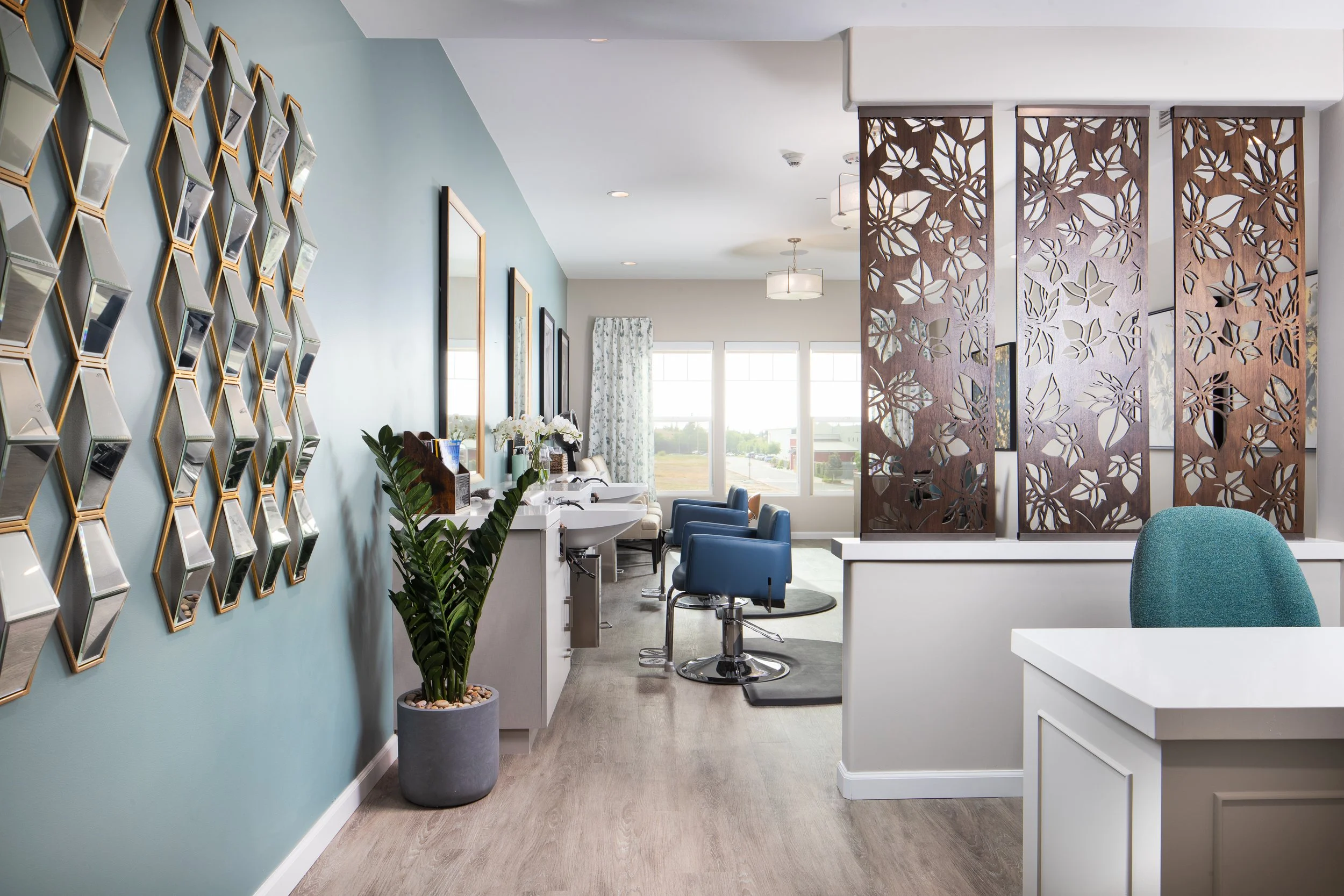 Salon with two sinks, blue chairs, decorative wall with hexagonal mirrors, large window with curtains, wooden room divider, and framed wall art.