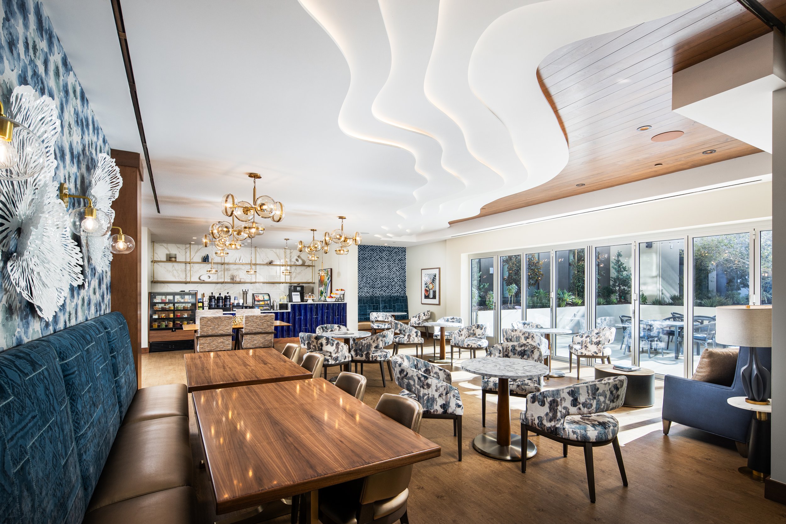 Bright, modern lounge or restaurant interior with wooden furniture, patterned chairs, large windows, and unique ceiling design with curved lighting.