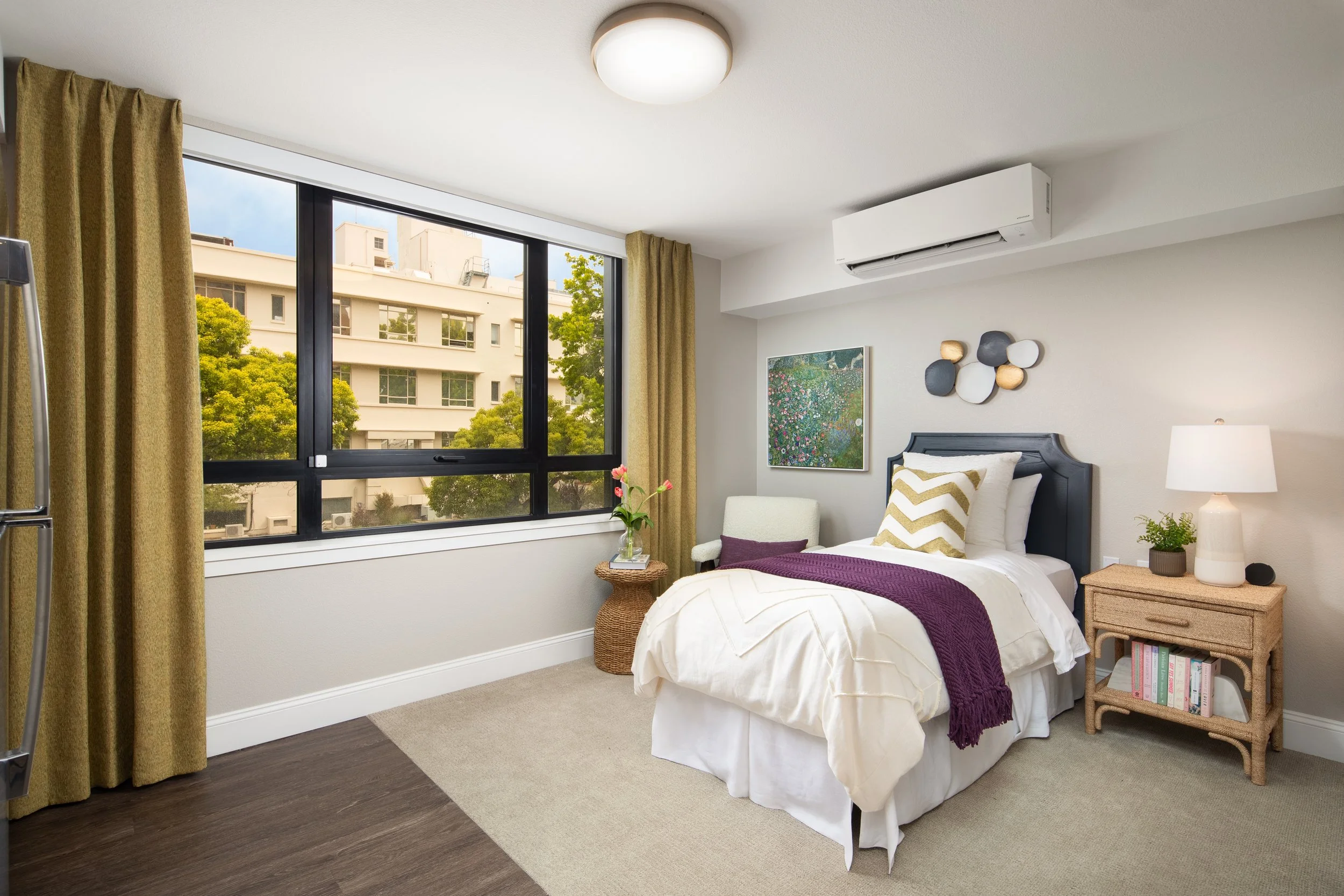 A bedroom with a large window showing trees and buildings outside, a bed with white bedding and a purple throw, a bedside table with a lamp, books, and a small plant, a wicker basket with a chair, and wall art.