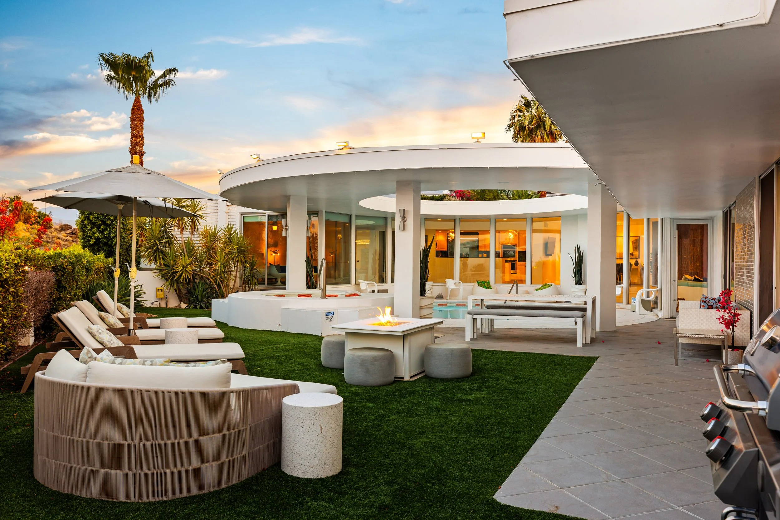 Modern backyard patio at sunset with lounge chairs, a fire pit, hot tub, glass-walled house, and outdoor kitchen with grill.