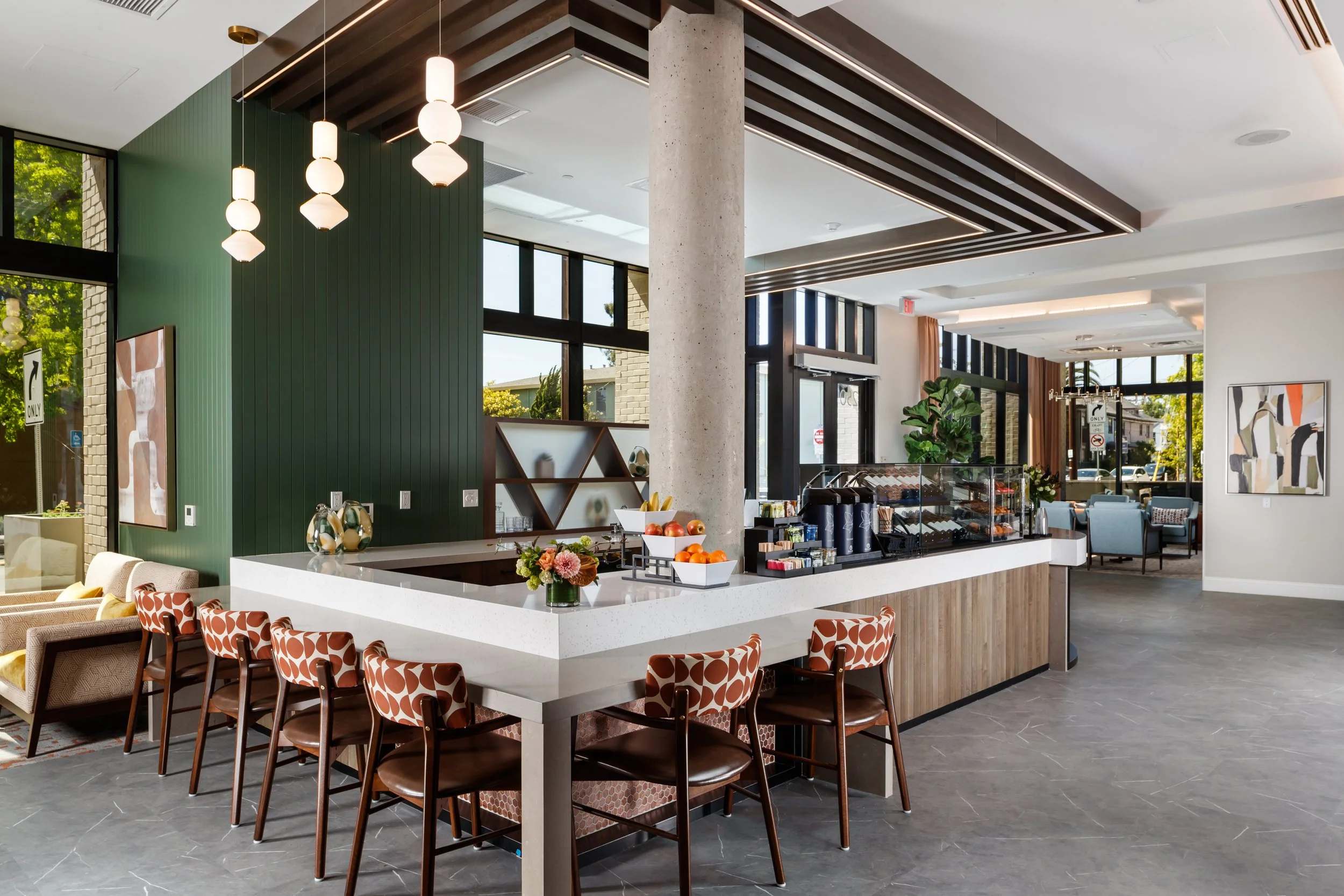 Modern cafe interior with a white stone counter, fruit display, flower arrangement, seating area with patterned chairs, large windows with natural light, and contemporary decor.