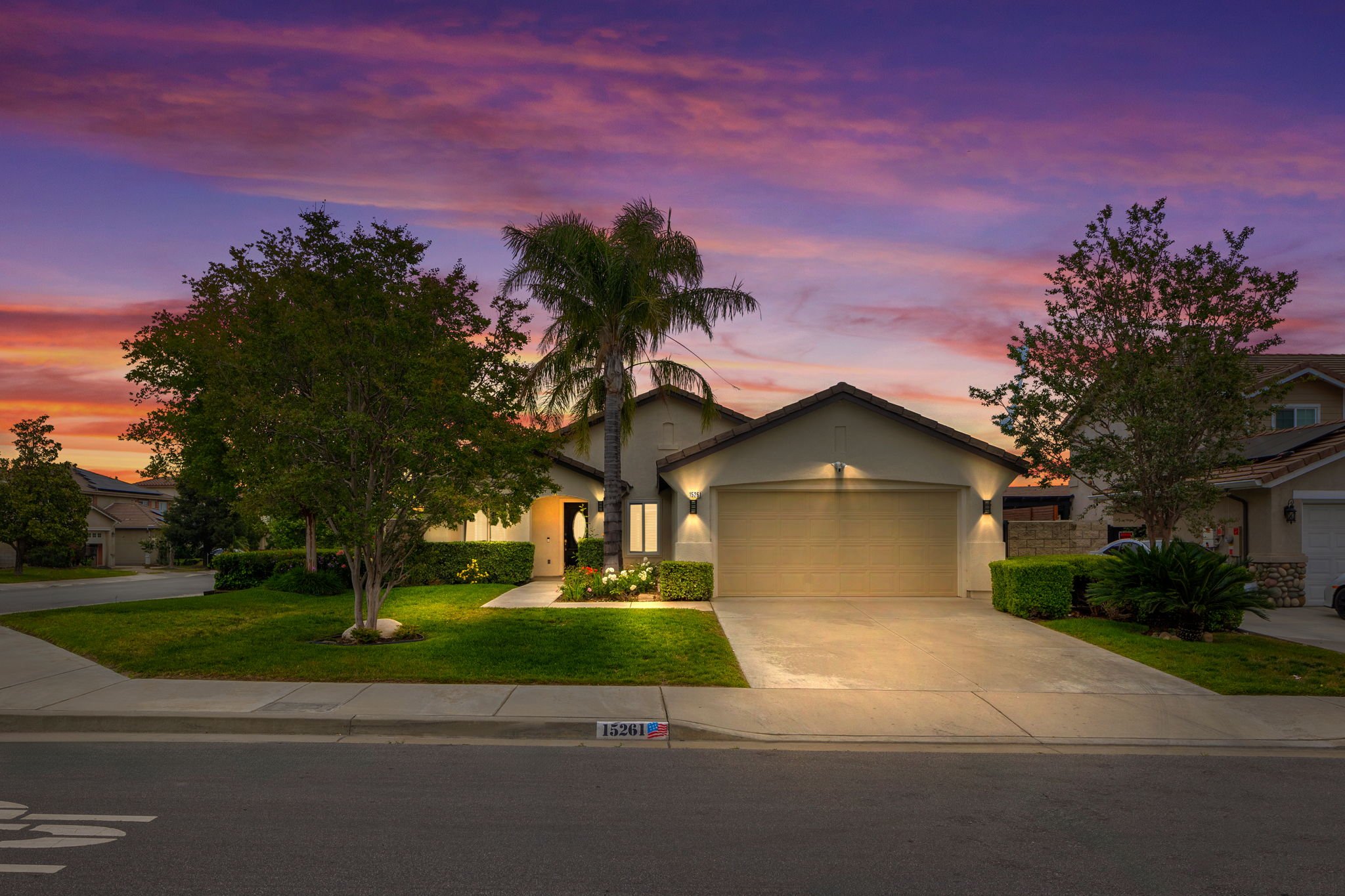 A suburban house with a beige garage door, surrounded by green trees and bushes, under a colorful sunset sky with purple, pink, and orange clouds.