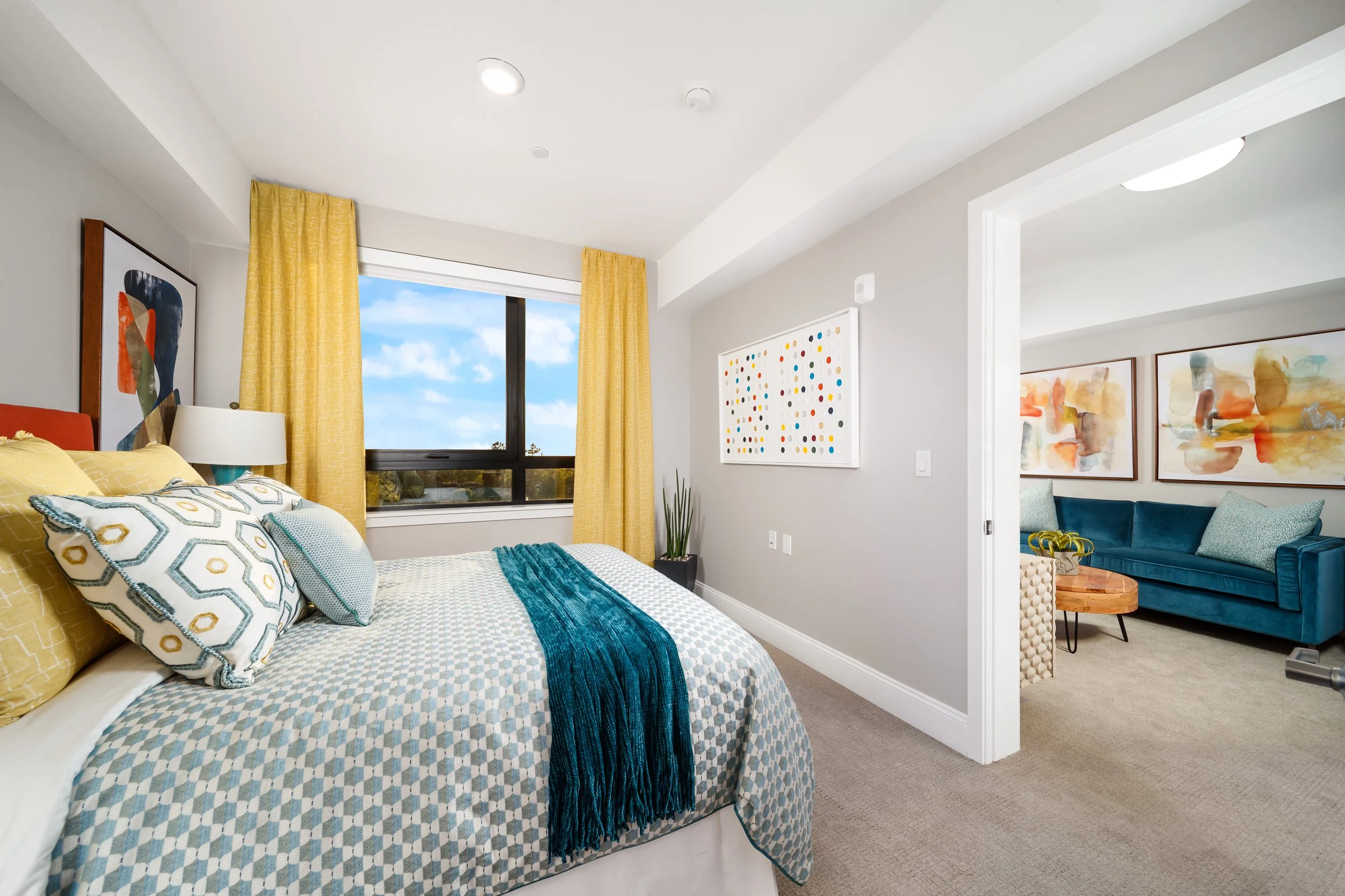 Freshly made bed with patterned bedding and throw pillows in a bright bedroom with yellow curtains, a large window, and artwork on the walls.