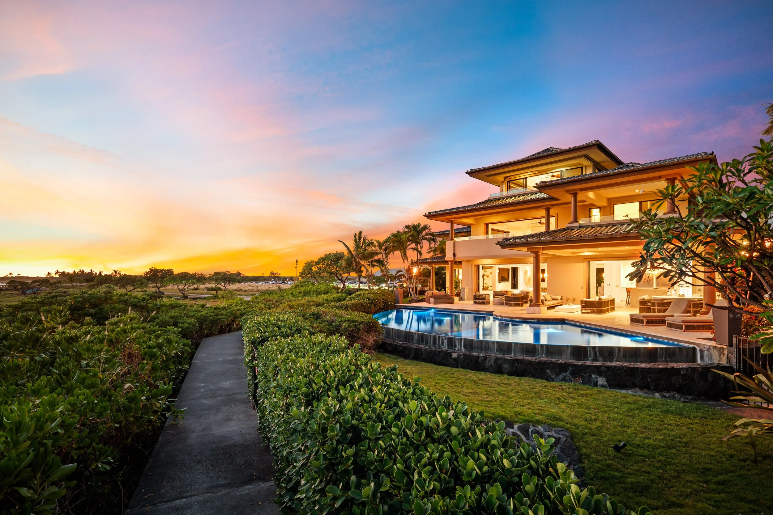 A modern house with large windows and outdoor seating area, a swimming pool, and lush green landscaping at sunset.