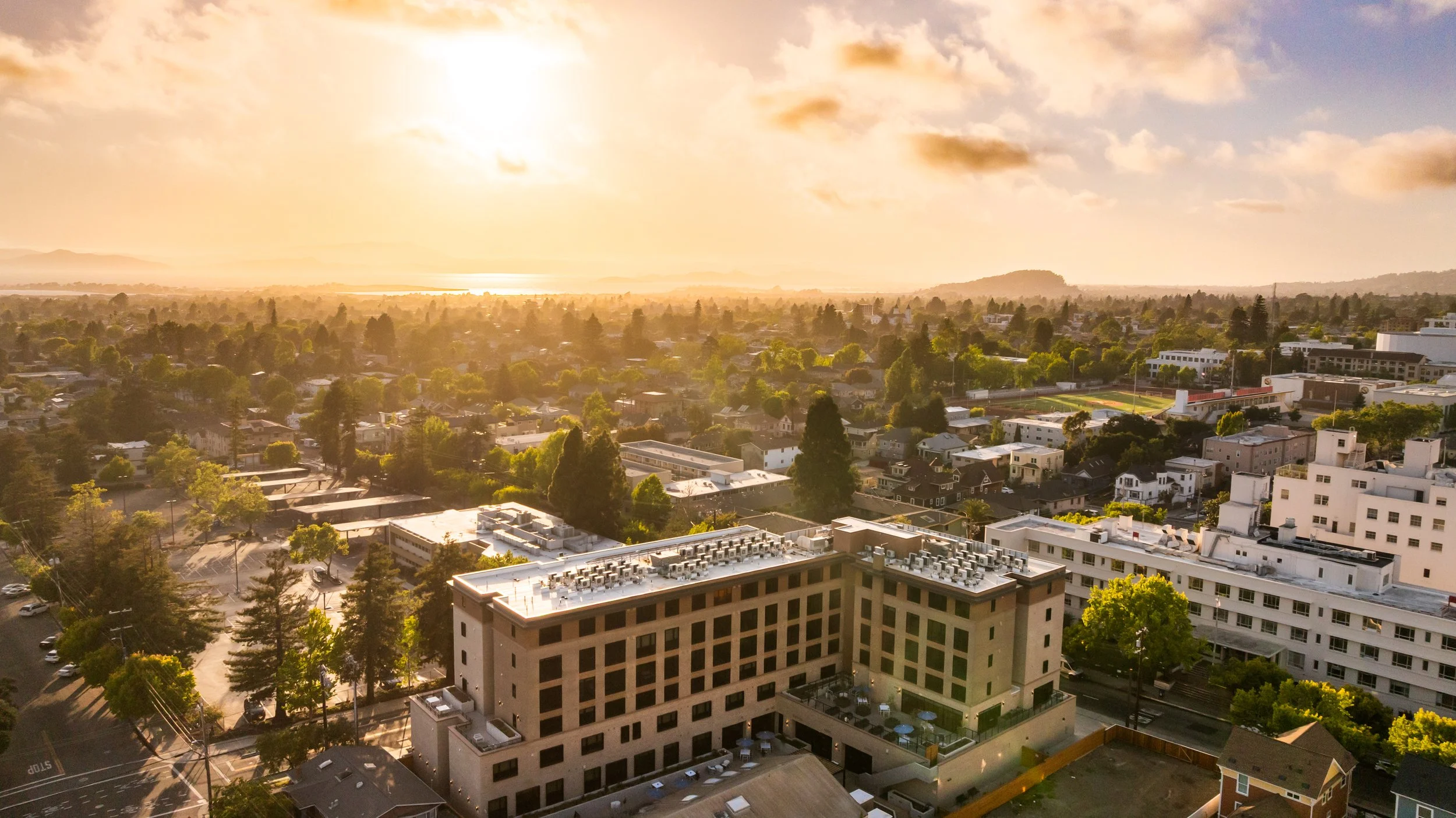 Aerial view of a cityscape at sunset with buildings, trees, and a horizon in the distance.