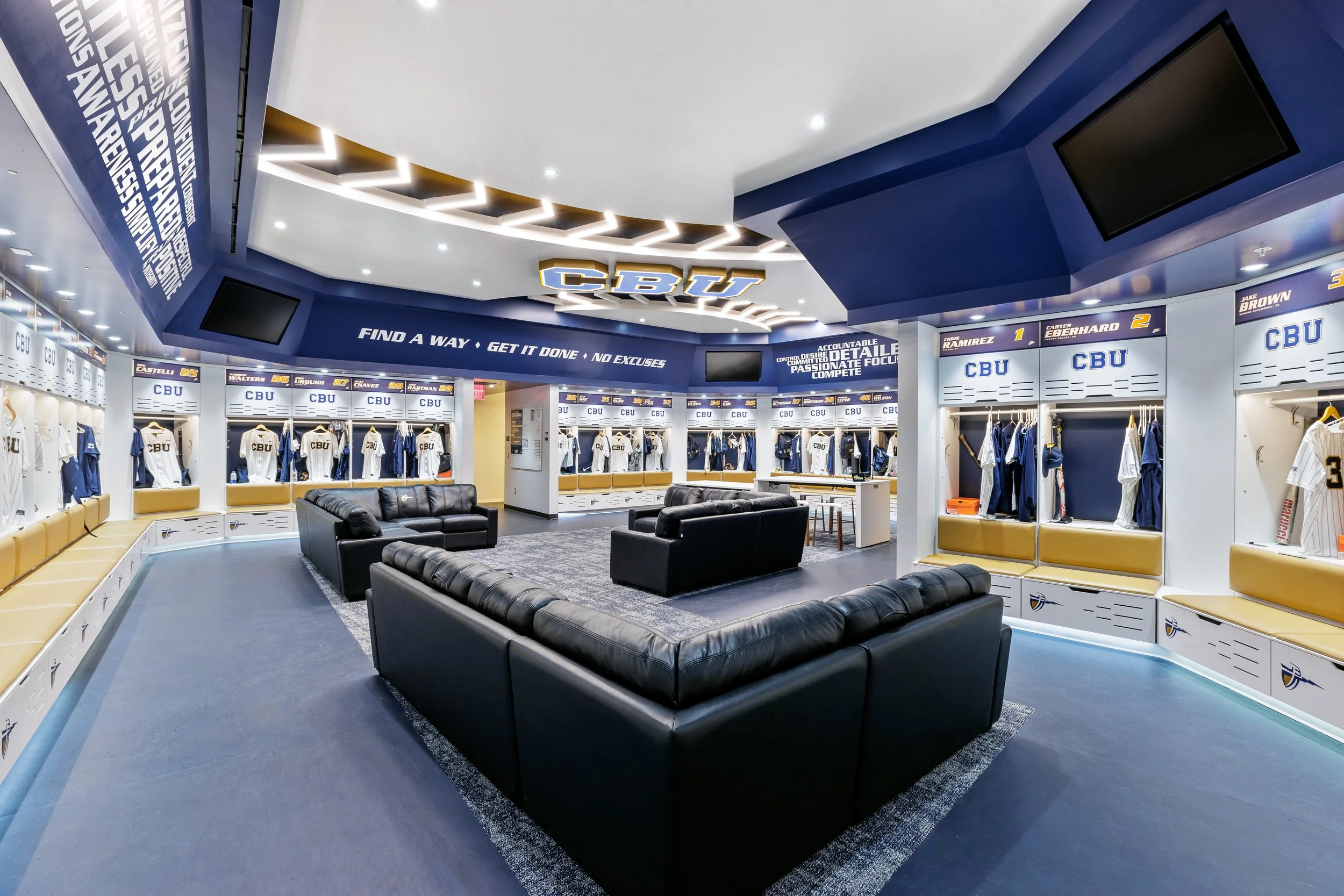 A locker room with black sofas in the center, surrounded by white lockers and team jerseys, with a blue and white decor and a large CBÚ sign on the ceiling.