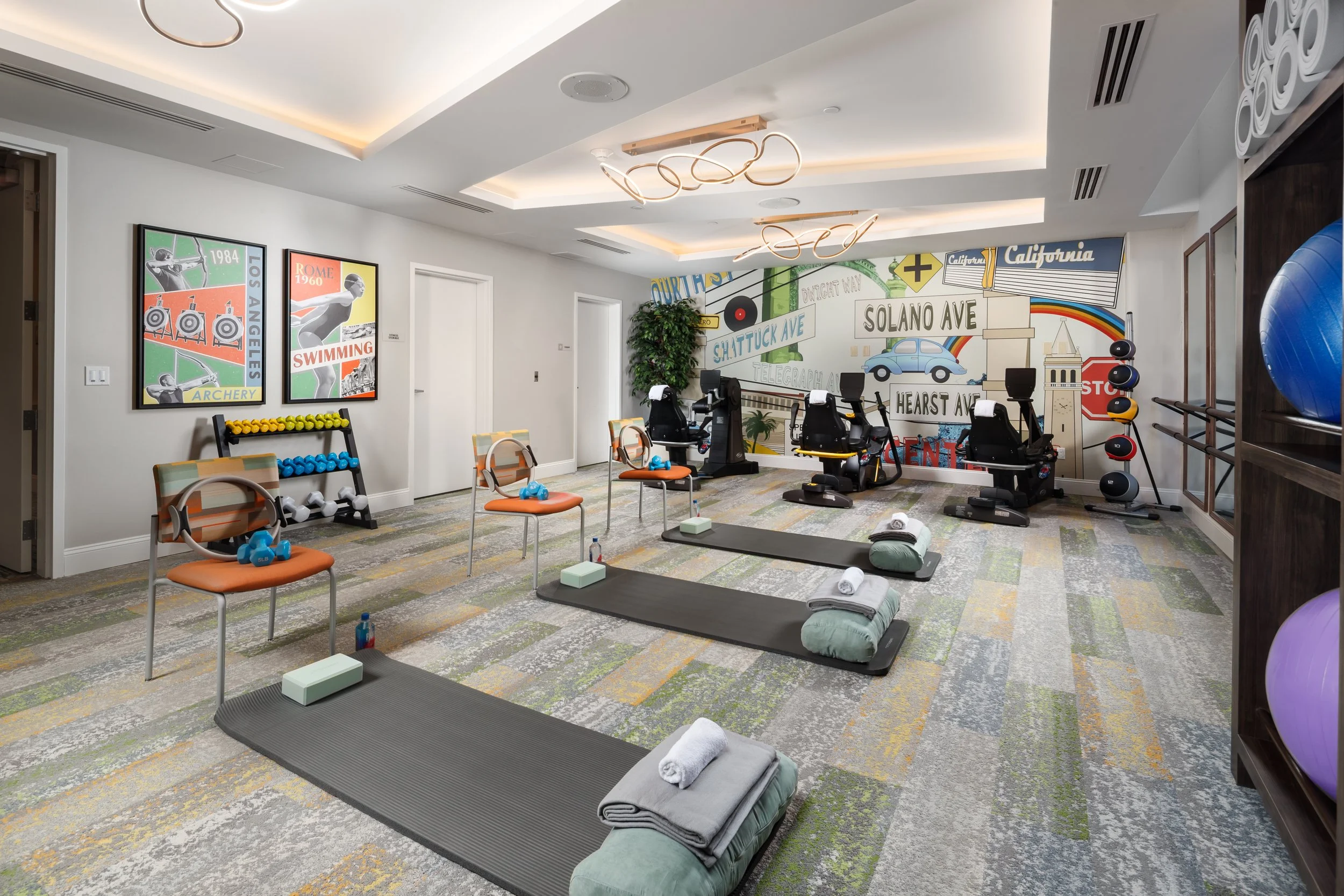 Indoor fitness room with exercise mats, resistance bands, dumbbells, exercise bikes, medicine balls, and a colorful mural on the wall depicting city street signs and landmarks.