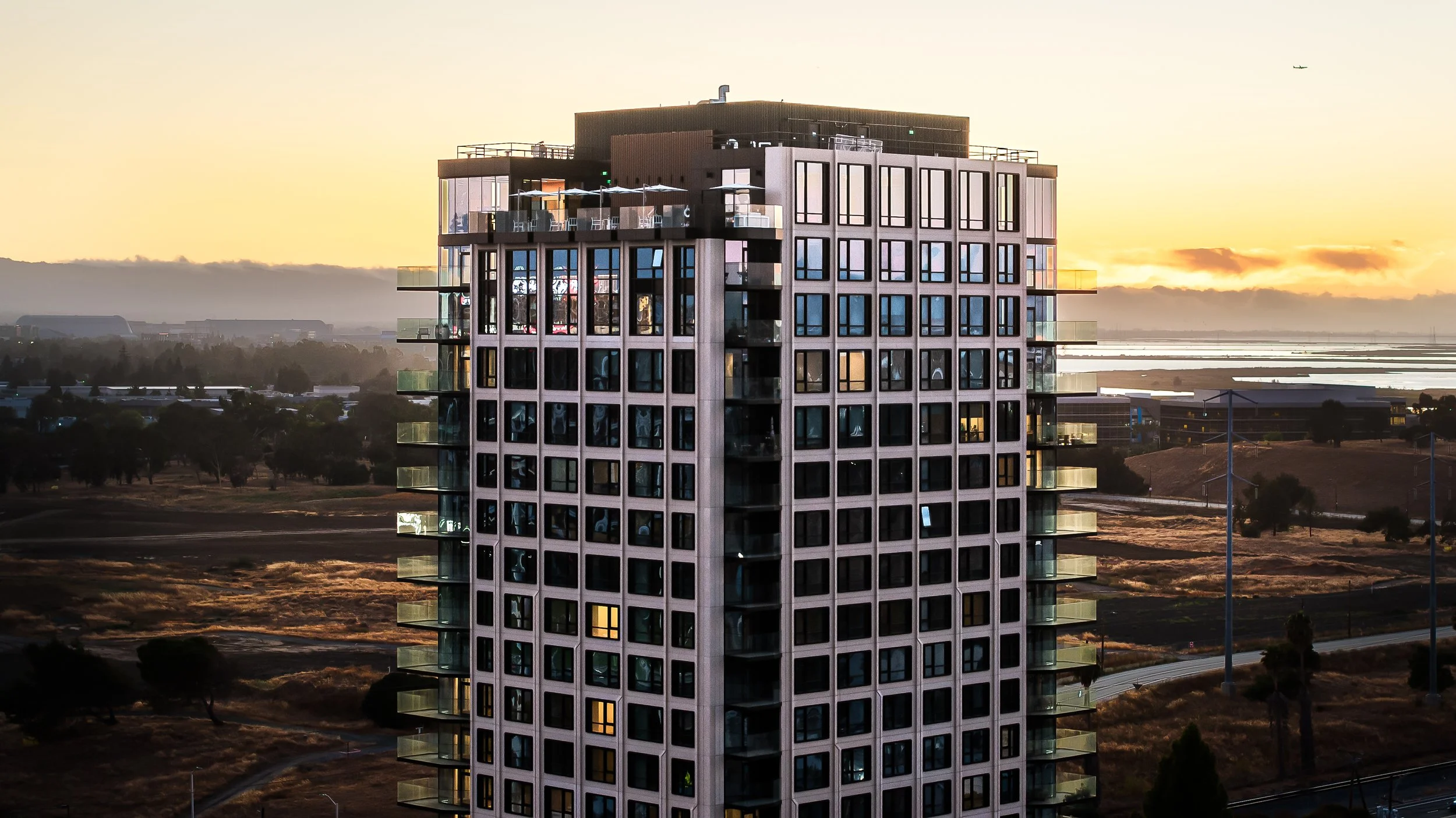High-rise apartment building with illuminated windows at sunset, surrounded by a landscape of trees, roads, and wind turbines.