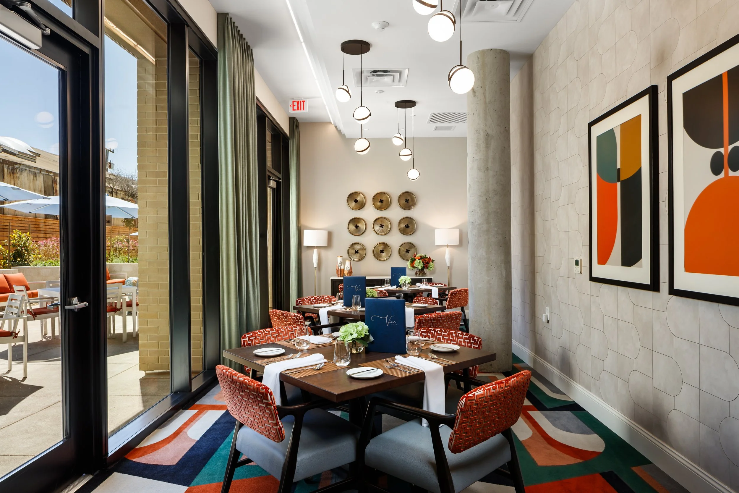 Elegant indoor dining area with a table set for four, featuring white tablecloths, plates, glasses, and napkins. The room has modern artwork on the walls, stylish lighting fixtures, and large windows with green curtains, offering a view of an outdoor