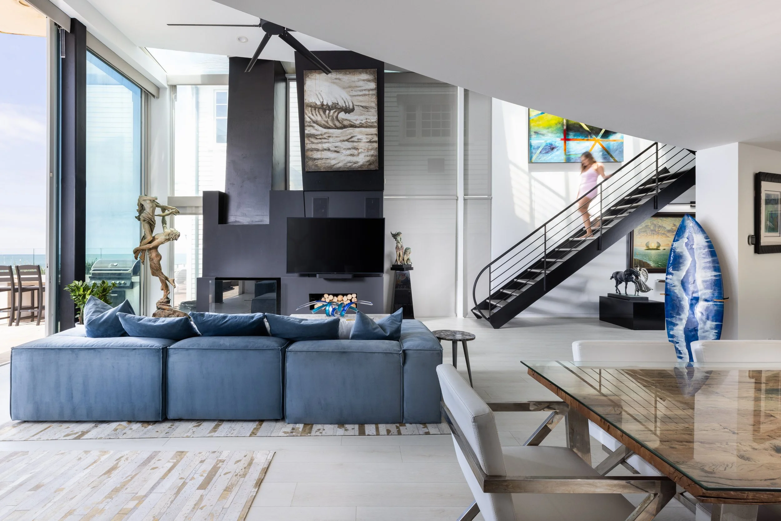 Modern living room with a large gray sofa, abstract paintings on black walls, a staircase with a person descending, and ocean view through large sliding glass doors.