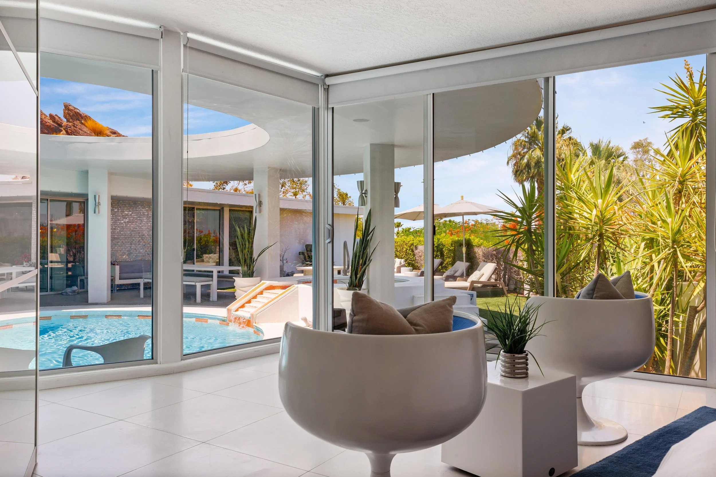 Living room with large glass windows overlooking pool area with lounge chairs, umbrellas, and lush greenery.