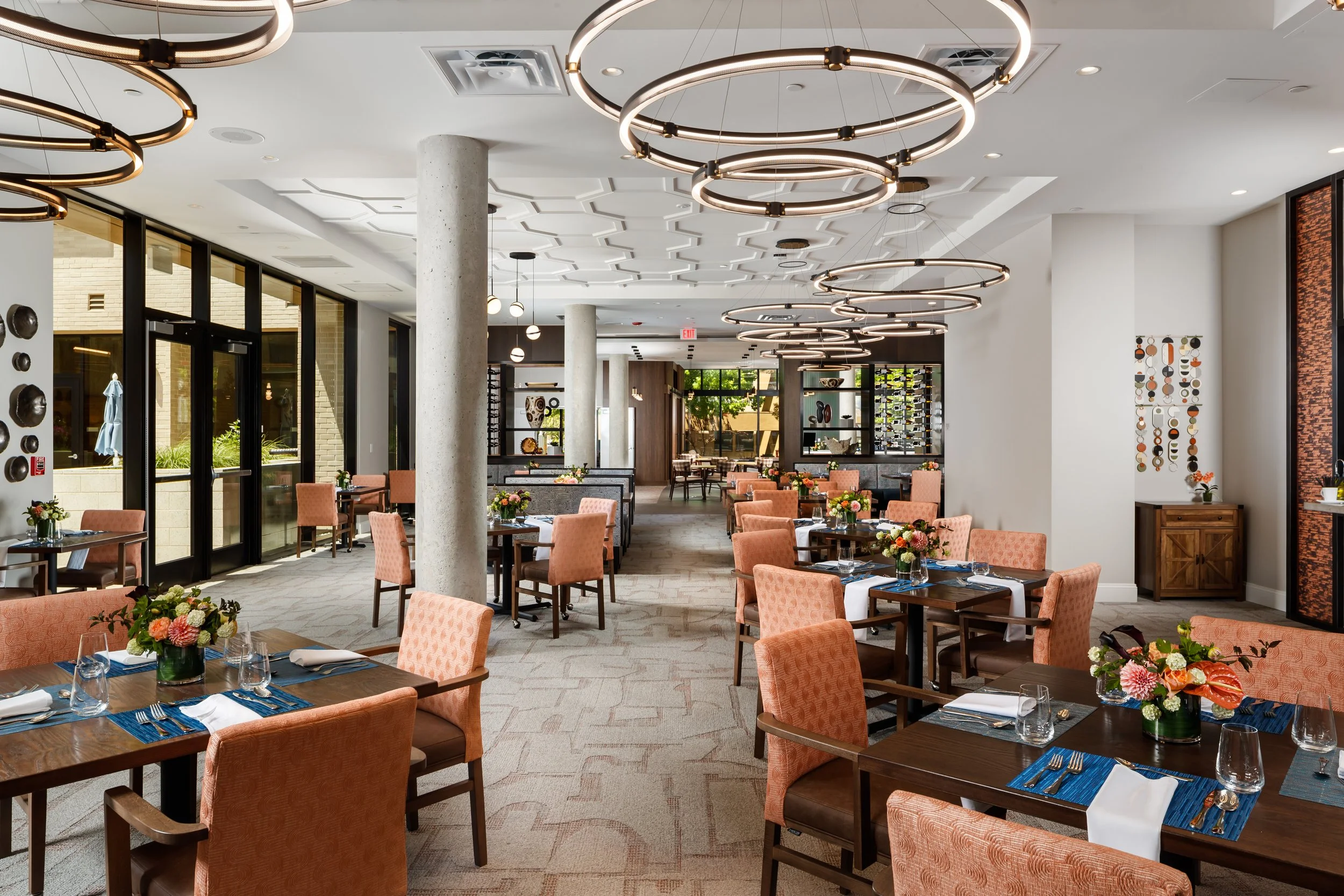 Elegant dining room with peach upholstered chairs, dark wood tables set with white napkins, cutlery, and glasses, floral centerpieces, large windows, modern circular chandelier lights, and decorative wall art.