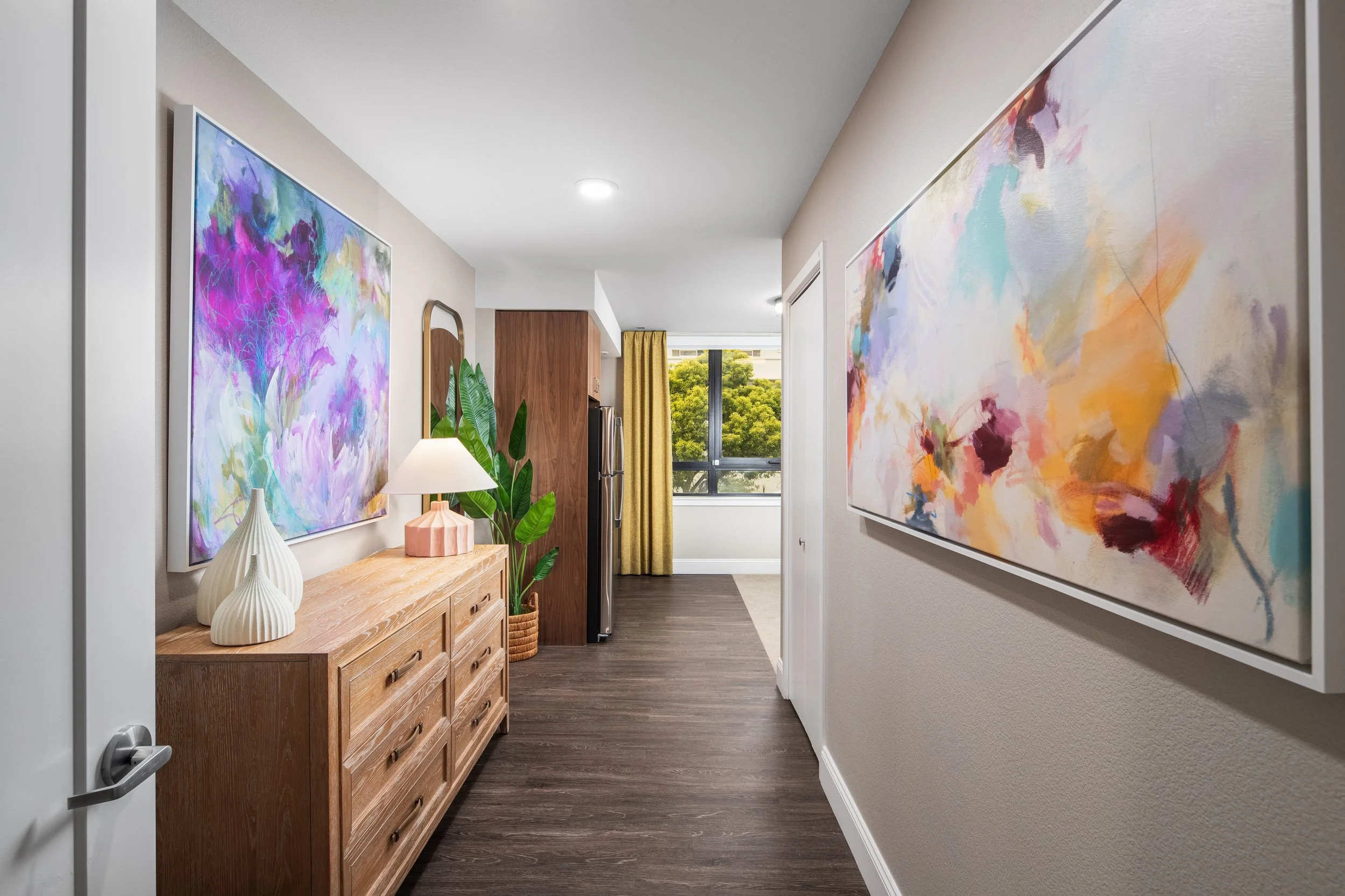 A hallway with two large abstract paintings on the walls, a wooden sideboard with decorative vases and a lamp, a potted plant, a small part of a refrigerator, and a window with yellow curtains showing greenery outside.