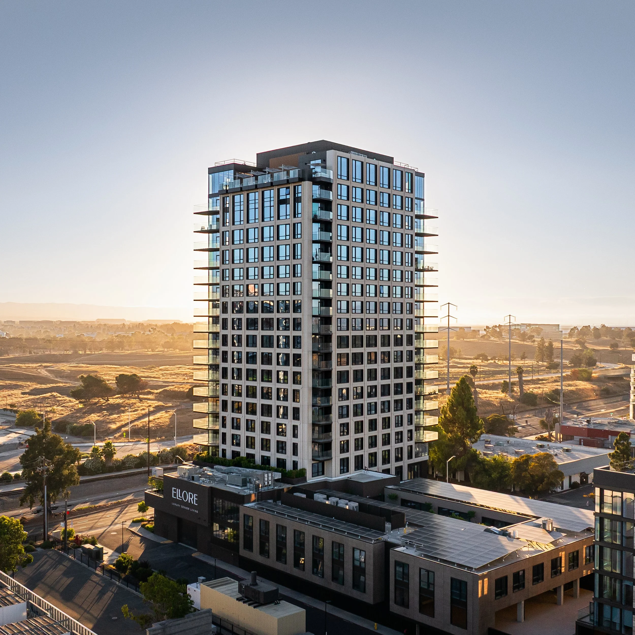 Tall modern residential or commercial building with many windows, balconies, and a sign that reads "Elore" at the bottom, set against a sunset sky with a semi-rural landscape in the background.
