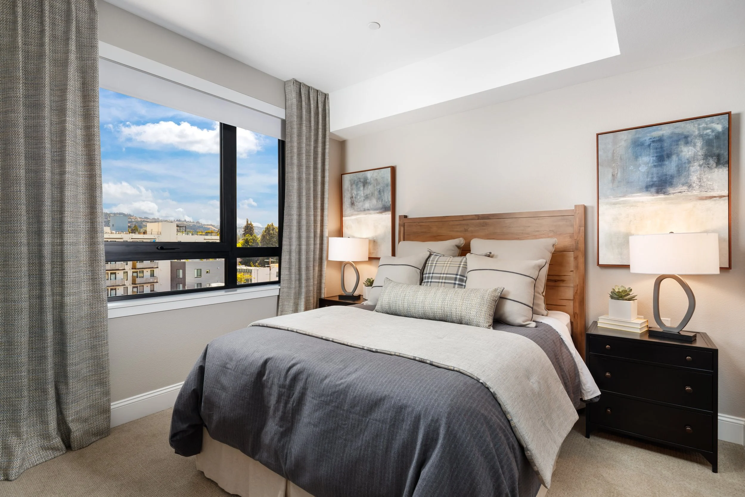 A bedroom with a large window looking out over city rooftops and hills, a wooden bed with multiple pillows, and nightstands with lamps and decorative items.