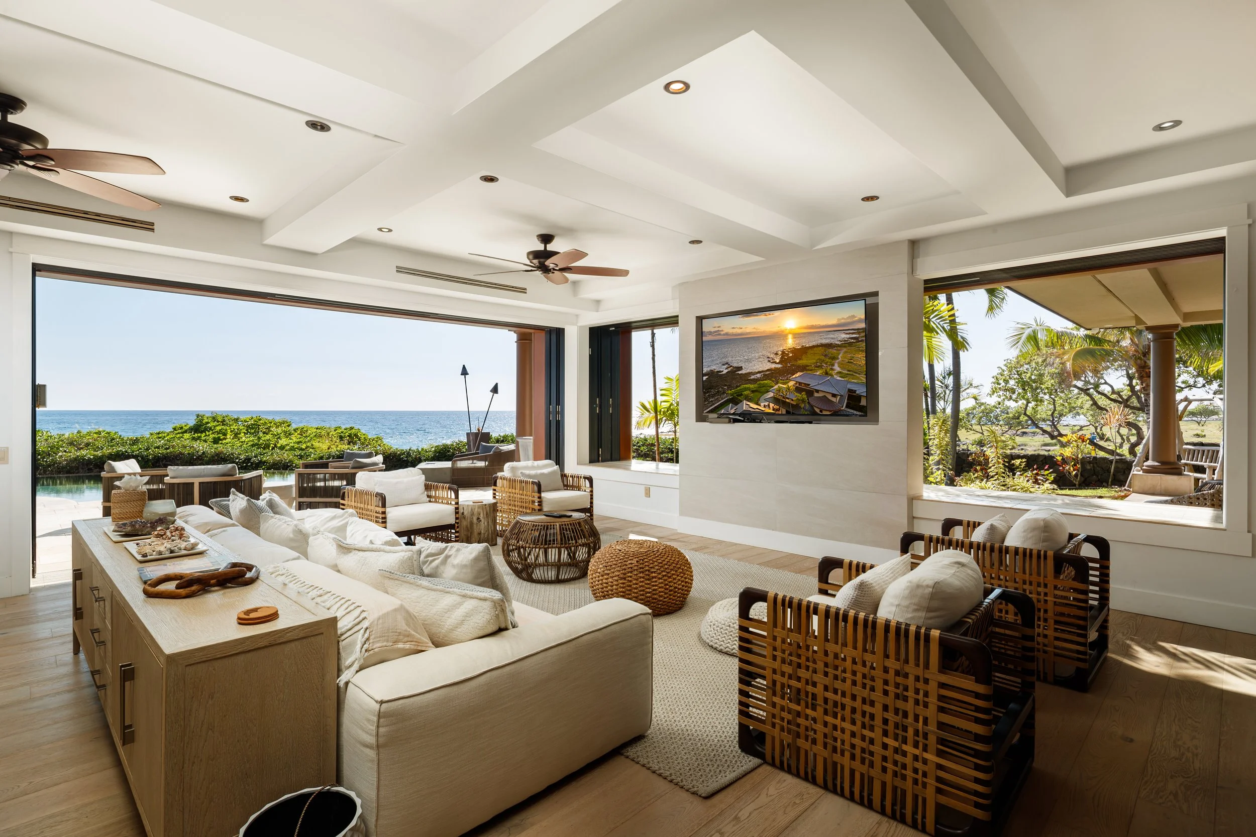 Living room with large windows showing ocean and garden views, beige sofa, wicker chairs, flat-screen TV, and wooden flooring.