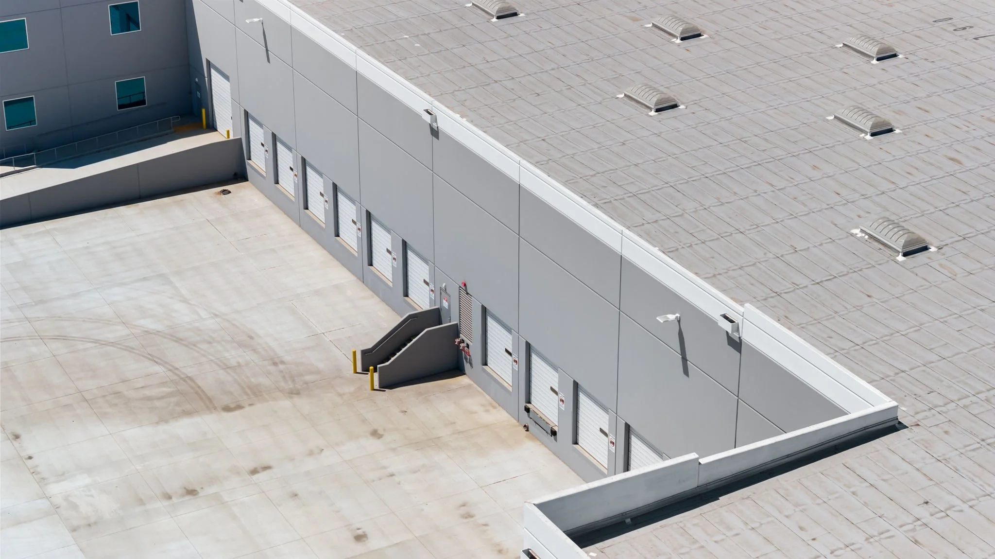 Aerial view of a commercial building's rooftop and loading dock area with multiple closed loading bay doors, stairs, and smoke vents on the roof.