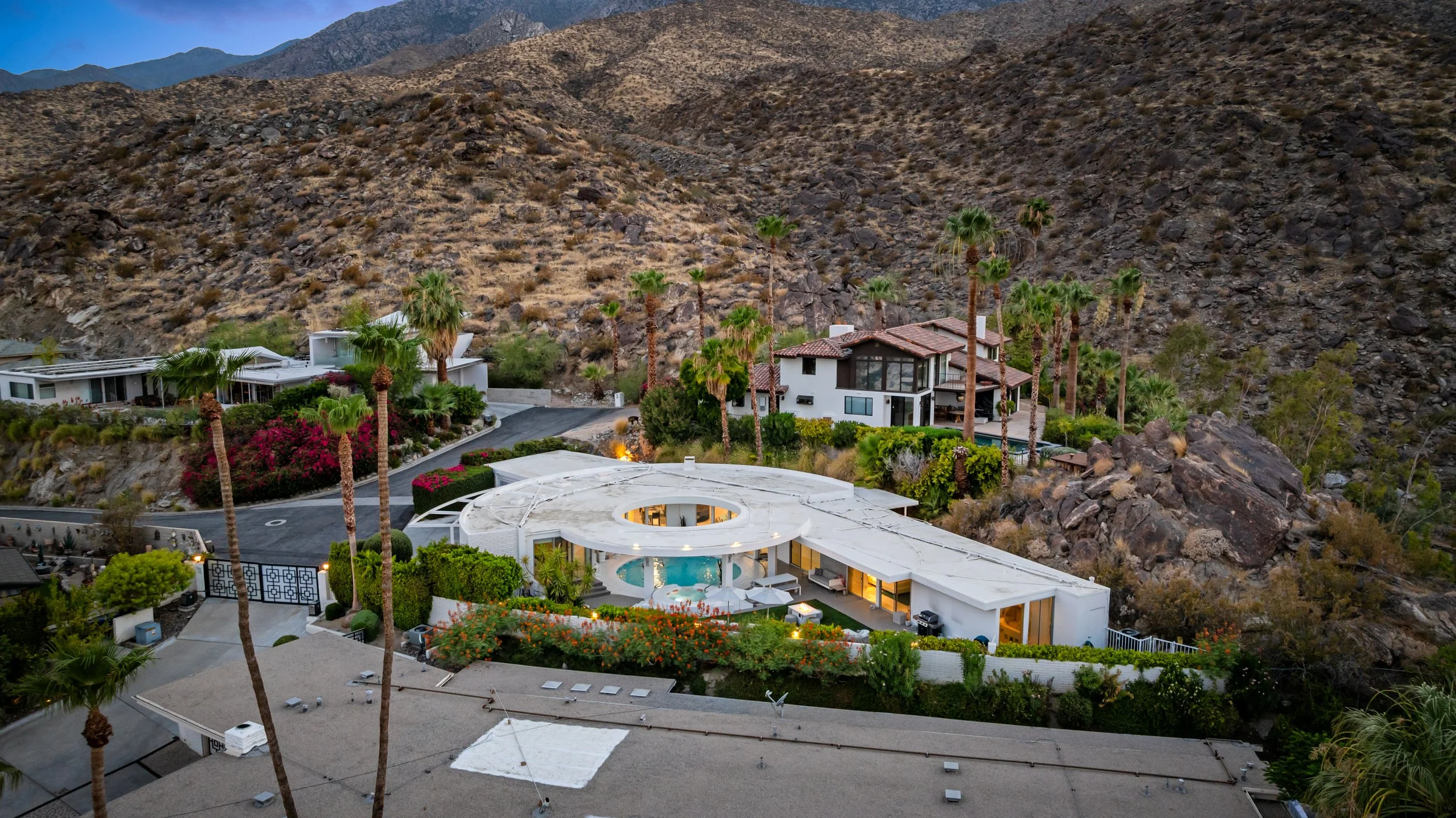 Aerial view of a modern white house with a pool, surrounded by palm trees and greenery, set on a hillside with rocky terrain and neighboring homes.