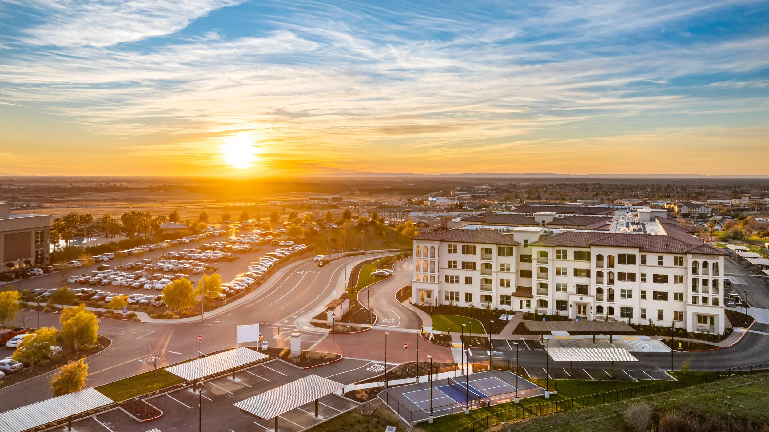 An aerial view of a modern apartment complex at sunset, with parking lots, tennis courts, and surrounding buildings under a colorful sky.