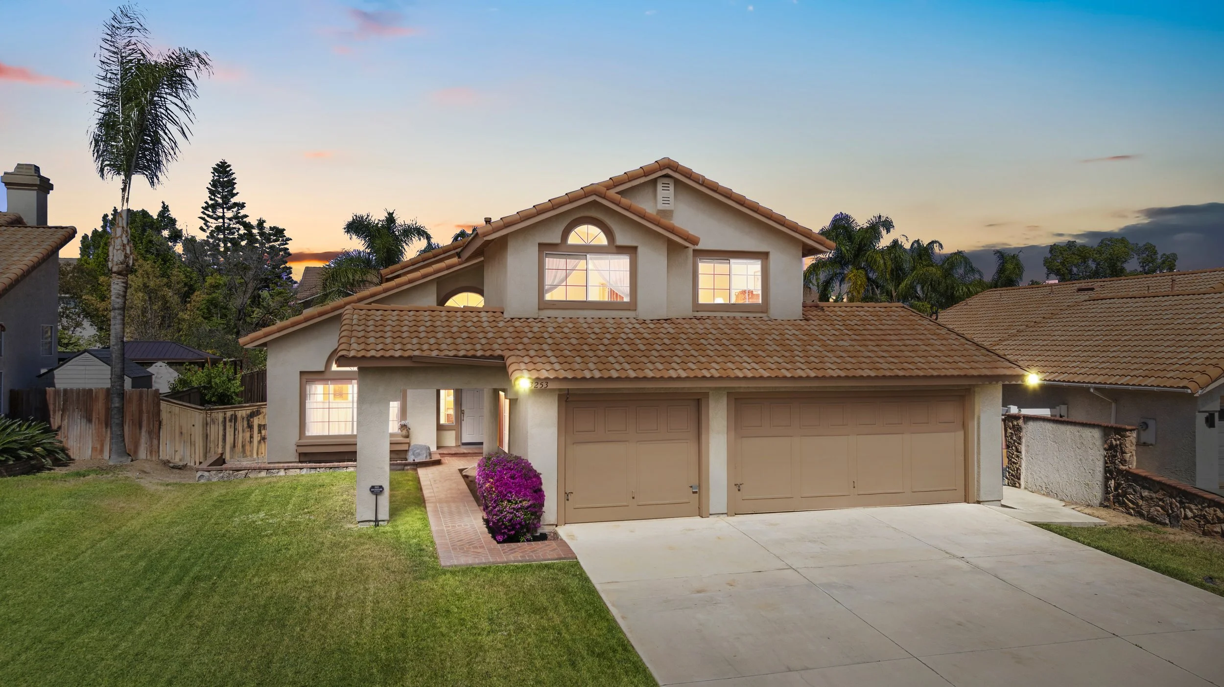Front view of a two-story house with a tan exterior, orange tile roof, three large windows, and a closed two-car garage. The yard has green grass, pinkish-purple flowers, and a brick walkway leading to the front door. The sky is at sunset with a colorful sky and trees in the background.