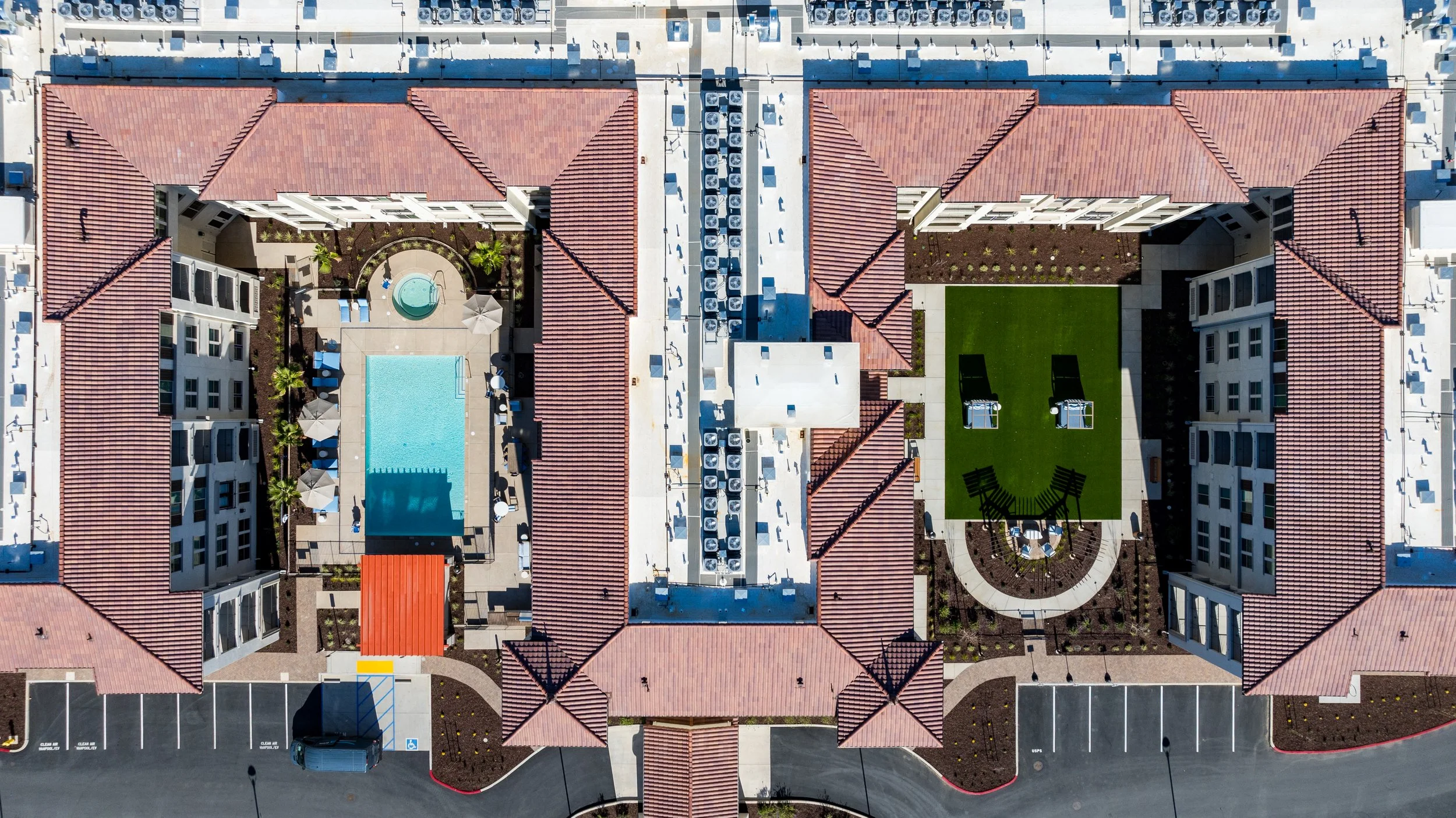 Aerial view of an apartment complex featuring a swimming pool, a hot tub, a lawn with two grills, and surrounding buildings with red tile roofs.