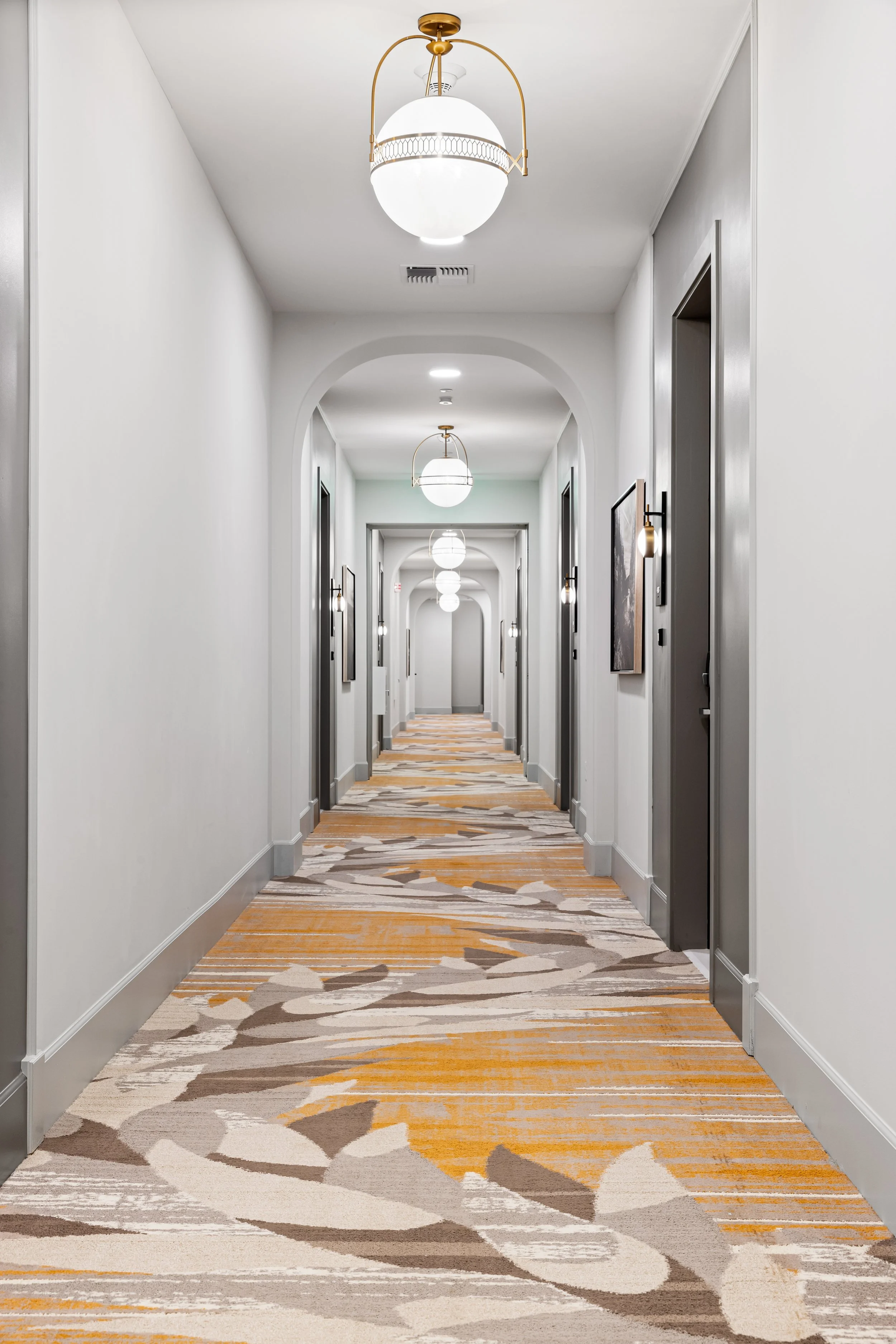 Long hotel hallway with white walls, gray door frames, and decorative wall-mounted lights on each side. The carpet has a modern pattern in shades of beige, gray, and orange. Hanging ceiling lamps with white globes and gold fixtures line the hallway.