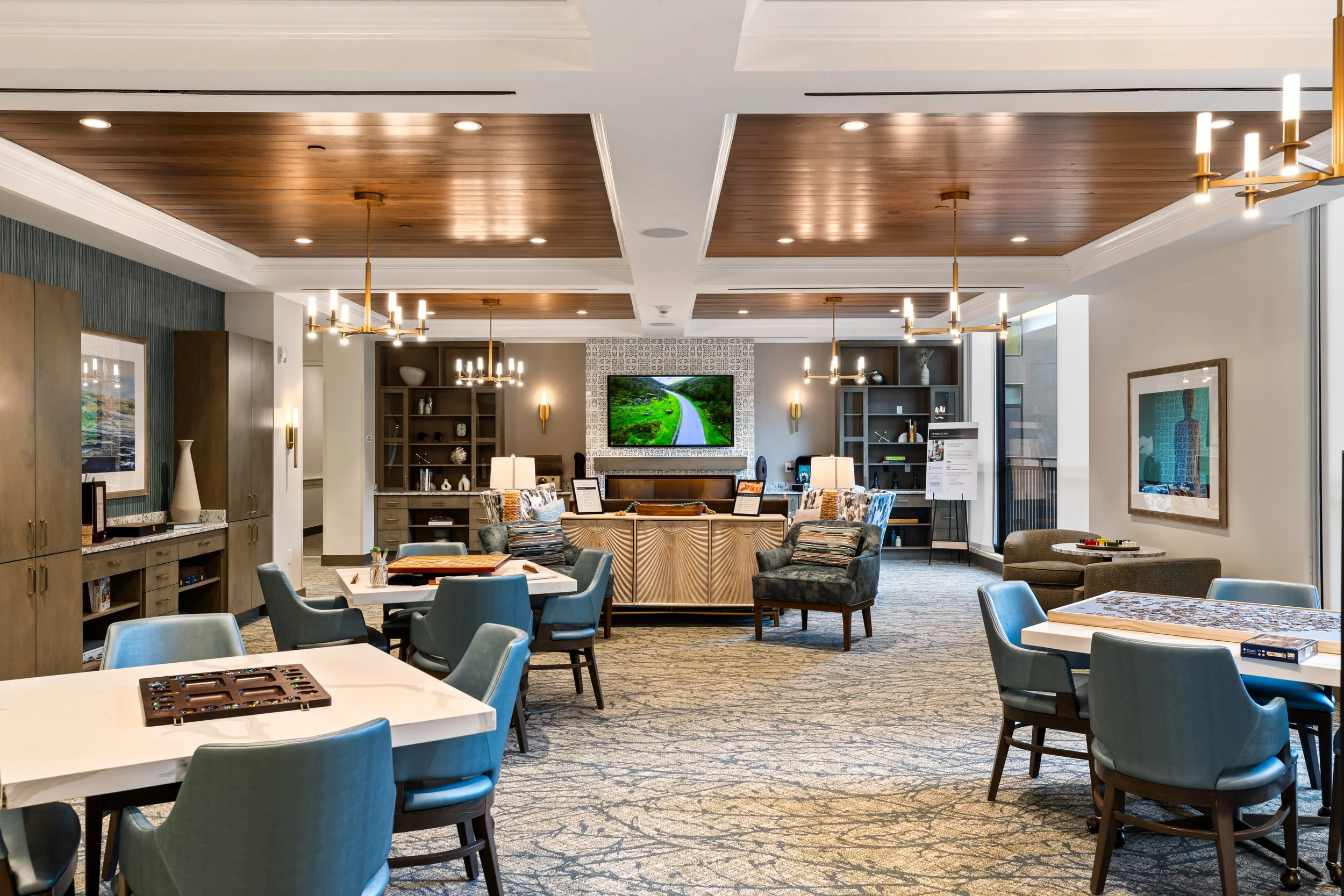 Interior of a modern lounge area with tables, chairs, bookshelves, a large wall-mounted television displaying a green landscape, and decorative lighting fixtures