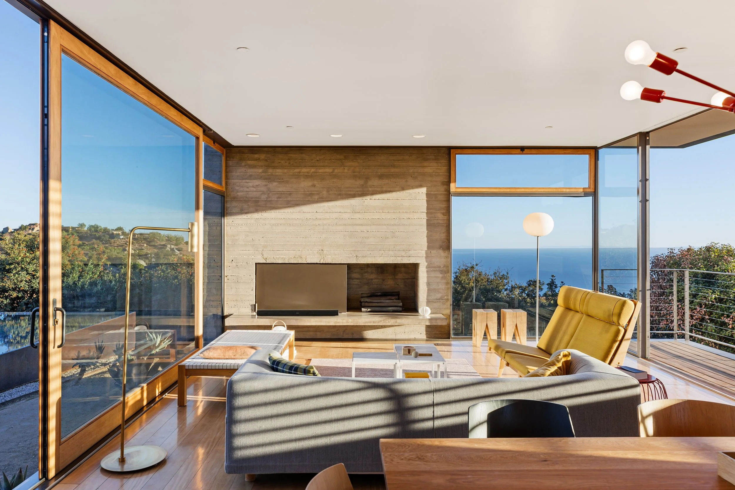 Modern living room with large glass windows showing ocean view, yellow lounge chair, gray sofa, white coffee table, wood accents, and a fireplace.