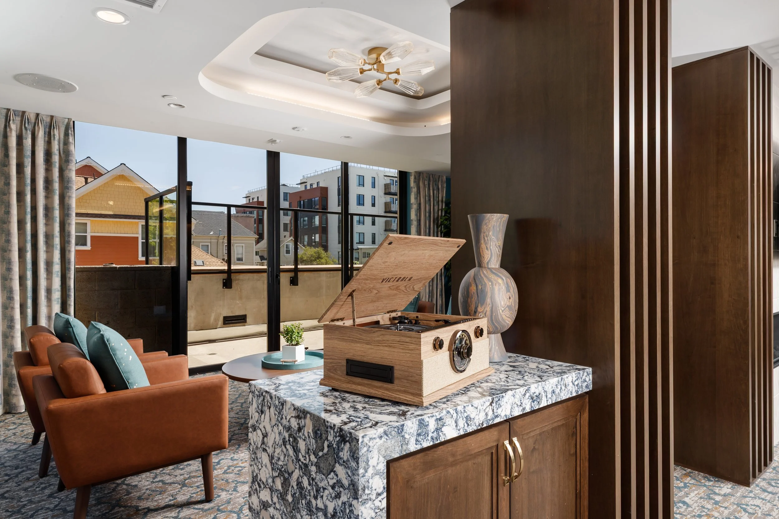 Living room with large windows, curtains, beige couches with teal pillows, a round table with a plant, a wooden record player on a marble countertop, and a decorative vase. The ceiling has a modern chandelier and recessed lighting.