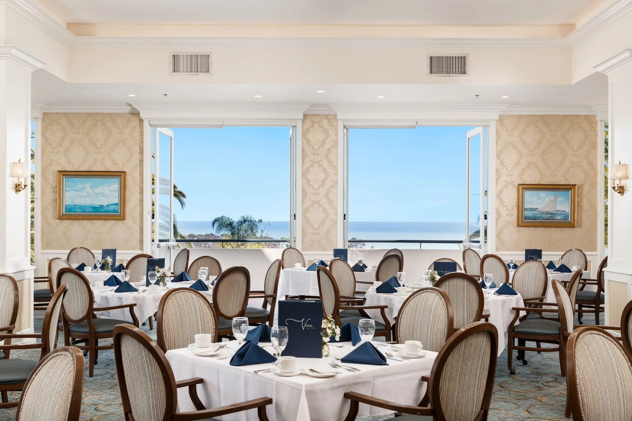 Elegant banquet room with round tables covered in white tablecloths, set with glassware and navy blue napkins, overlooking a view of the ocean through large windows, with framed sailboat paintings on the walls.
