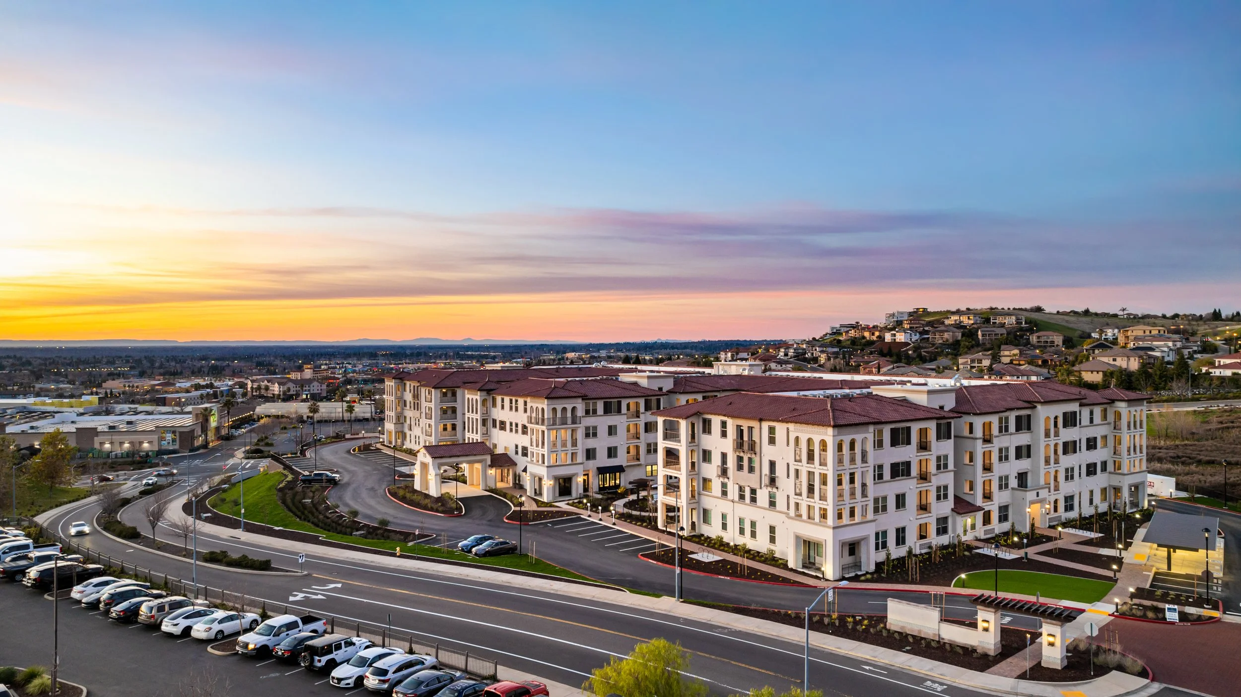 A modern residential apartment complex with multiple white buildings, red tile roofs, and a curved driveway at sunset, with parking lots, green lawns, and surrounding hills in the background.