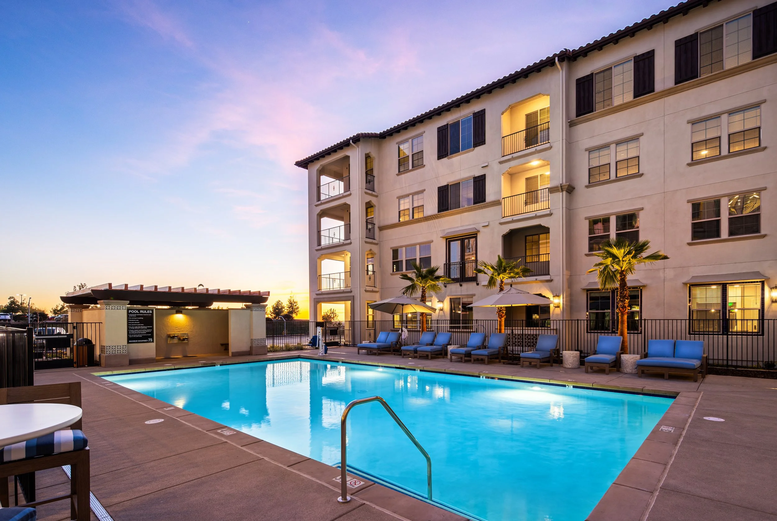 An outdoor swimming pool area at sunset, with lounge chairs, umbrellas, and a multi-story residential building in the background.