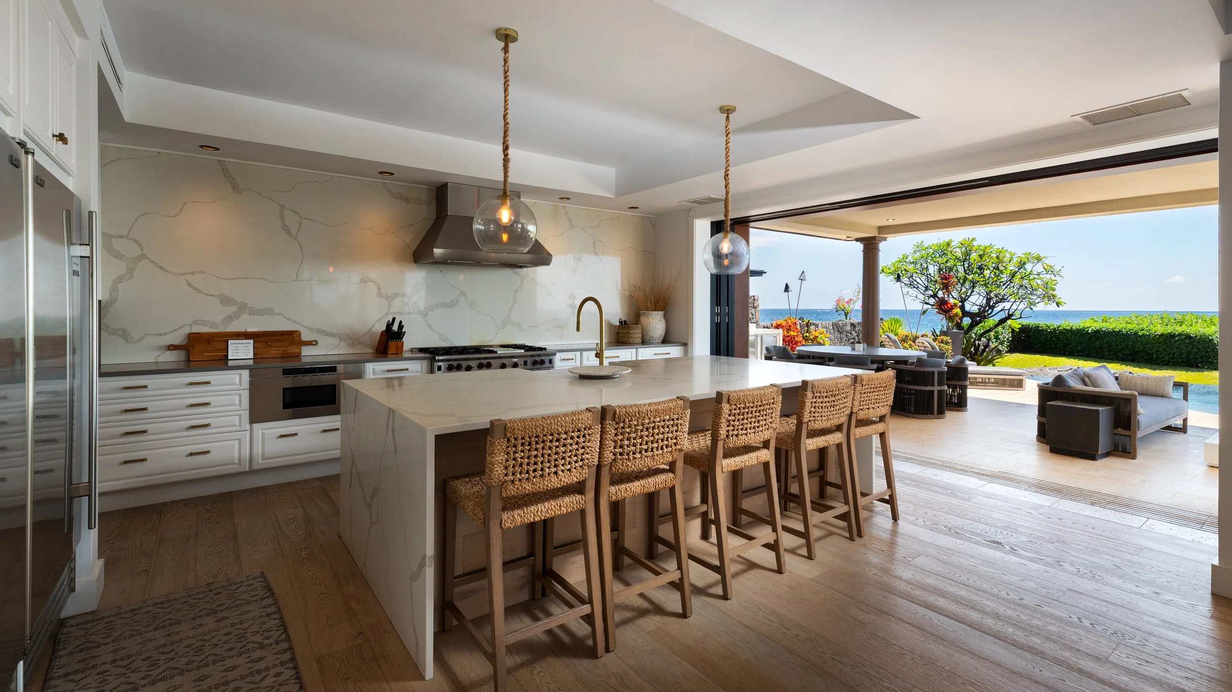 Open-concept kitchen with white cabinets, marble countertops, a large island with four woven bar stools, and a view of an outdoor patio with furniture and ocean scenery.