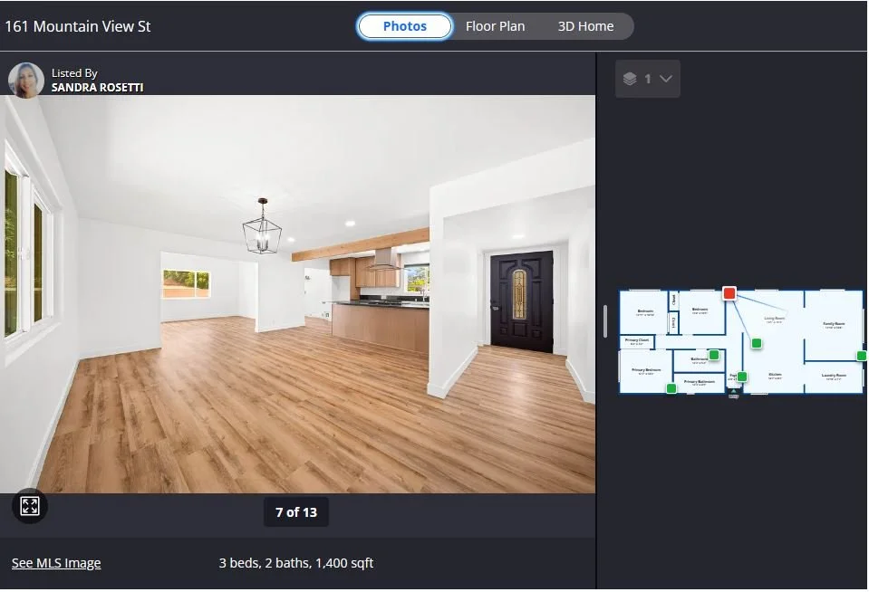 Interior view of a spacious, empty living room with hardwood floors, white walls, and large windows. There is a kitchen with wooden cabinets, a black countertop, and a geometric pendant light hanging from the ceiling near the open living space. A door with a decorative glass panel is visible on the right side.
