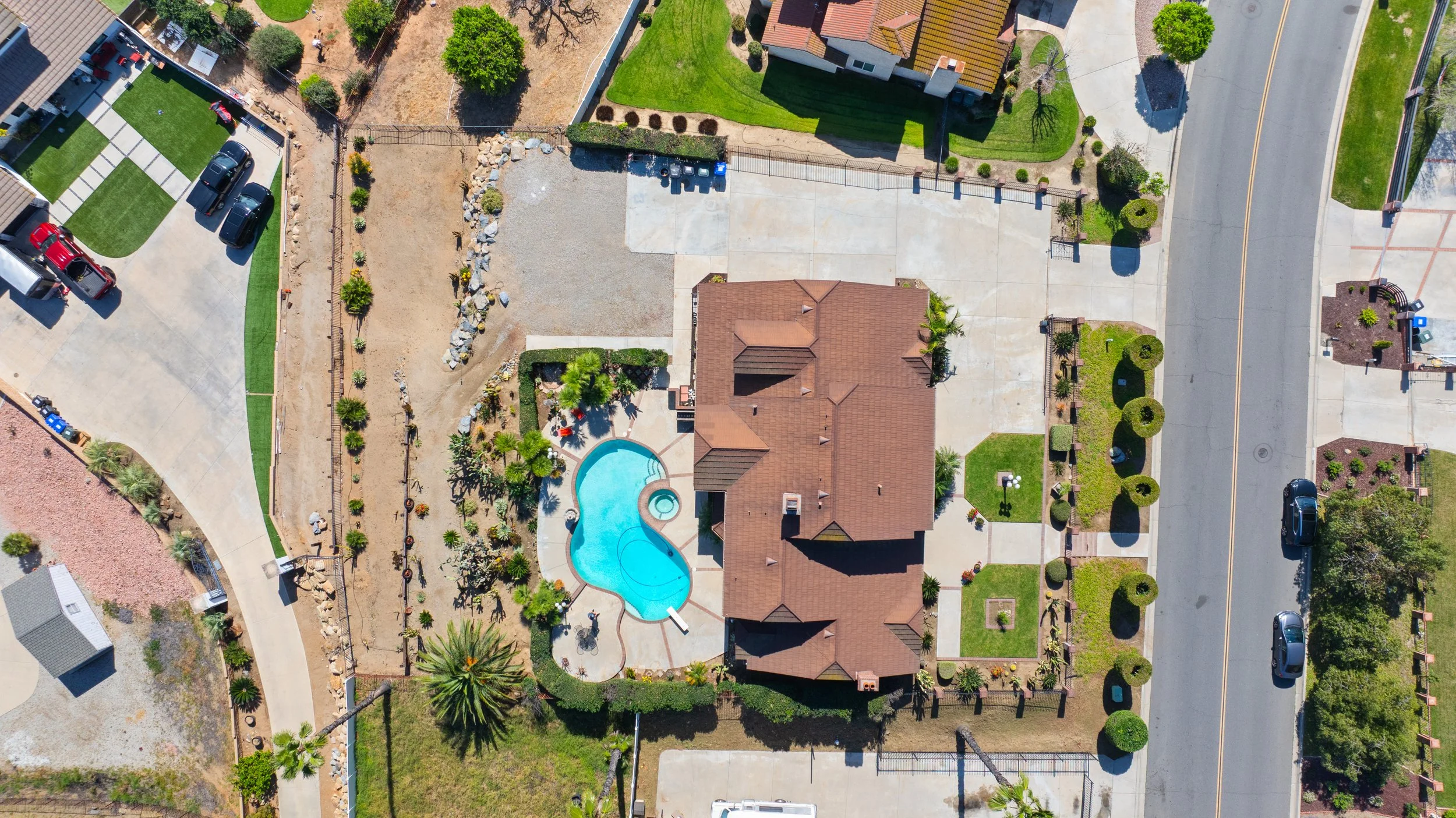 Aerial view of a residential backyard with a swimming pool, a house with a brown roof, a small yard, and a driveway. Surrounding lawns and trees are visible, along with neighboring houses and streets.