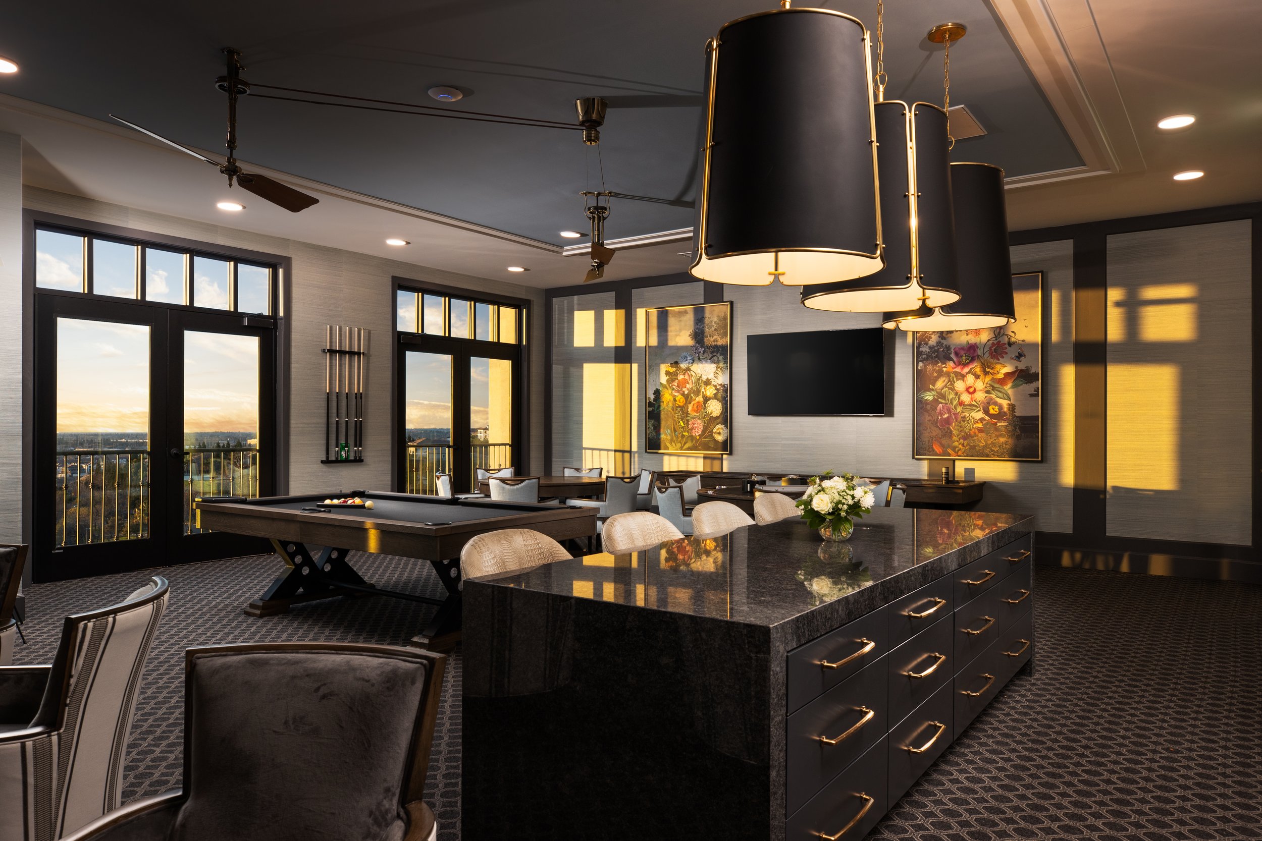 Elegant indoor lounge with large windows showing a sunset view, featuring a pool table, a black granite counter with gold handles, a flower arrangement, a mounted TV, and modern decor.