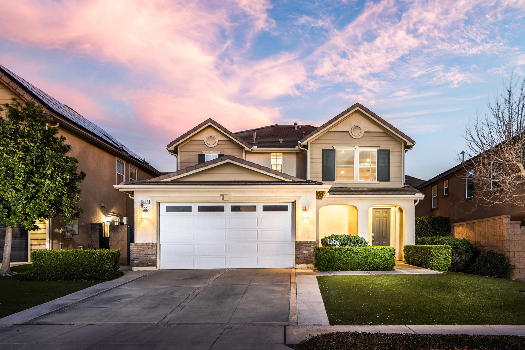 A two-story suburban house with a white garage door, beige siding, and black shutters, illuminated by exterior lights at dusk with a pastel pink and blue sky overhead.