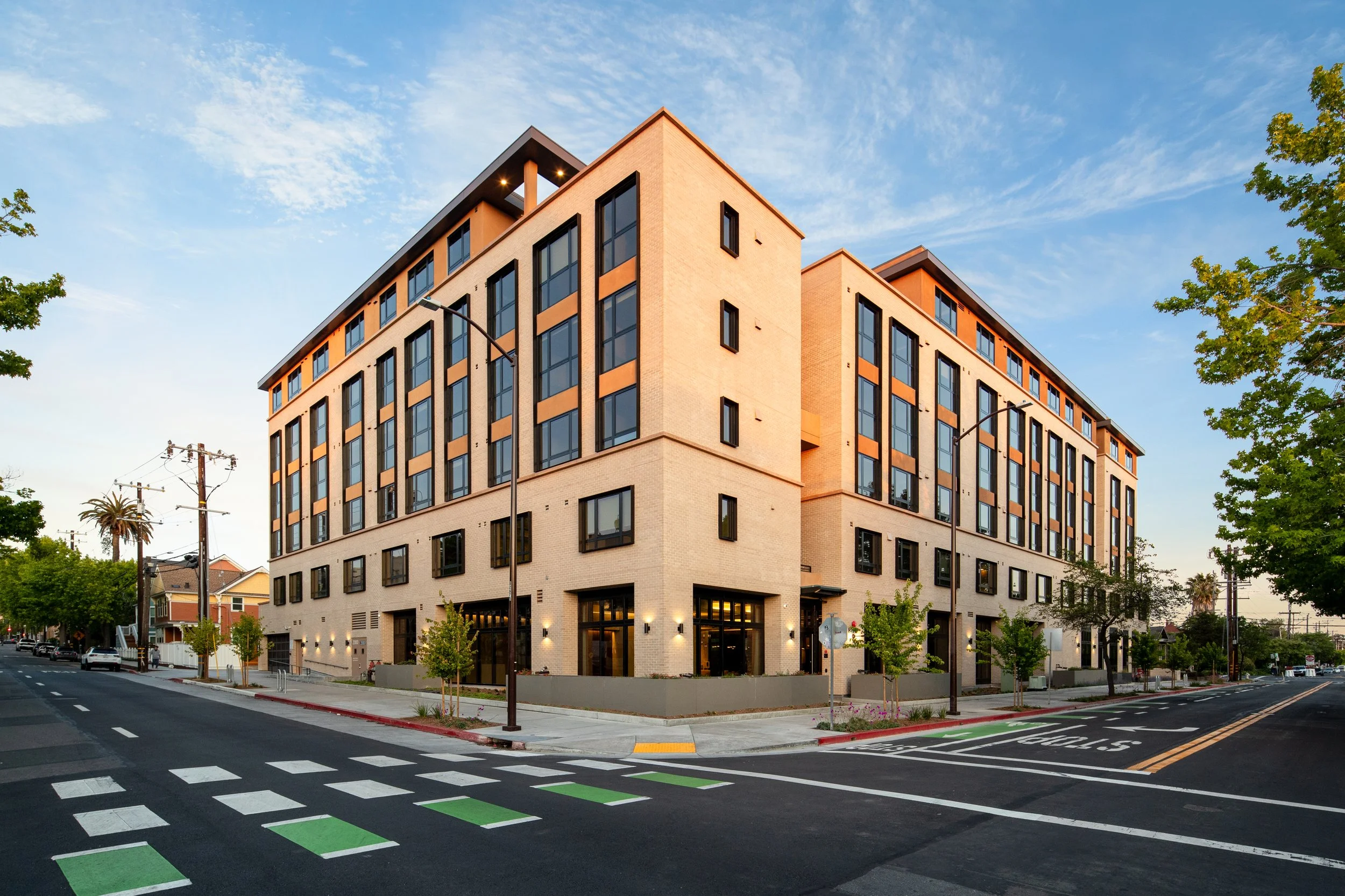 A modern multi-story residential building with large windows at the corner of a street during daylight, surrounded by trees and streetlights.